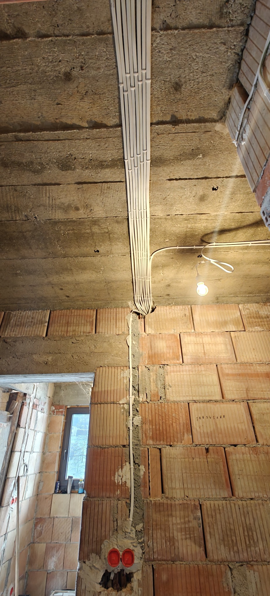 Wnętrze w trakcie prac: kable w korytkach na suficie, ściany z czerwonej cegły, widoczne puszki instalacyjne. Surowy stan, przygotowanie do dalszych prac wykończeniowych.