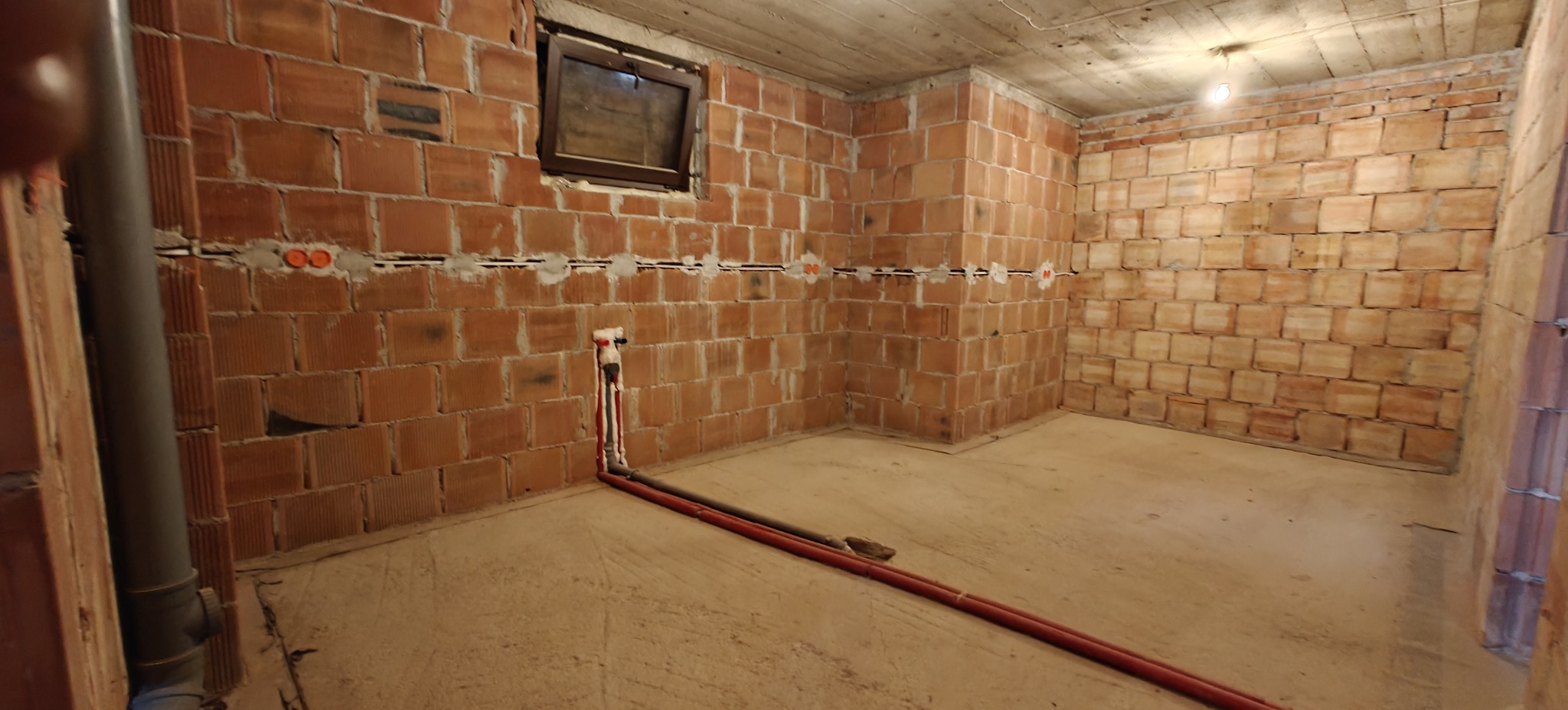 Surowy pokój z ceglanymi ścianami w trakcie prac instalacyjnych. Widoczne rury i puszki elektryczne w ścianach. Okno w górnej części ściany. Stan surowy, przygotowanie do wykończenia.