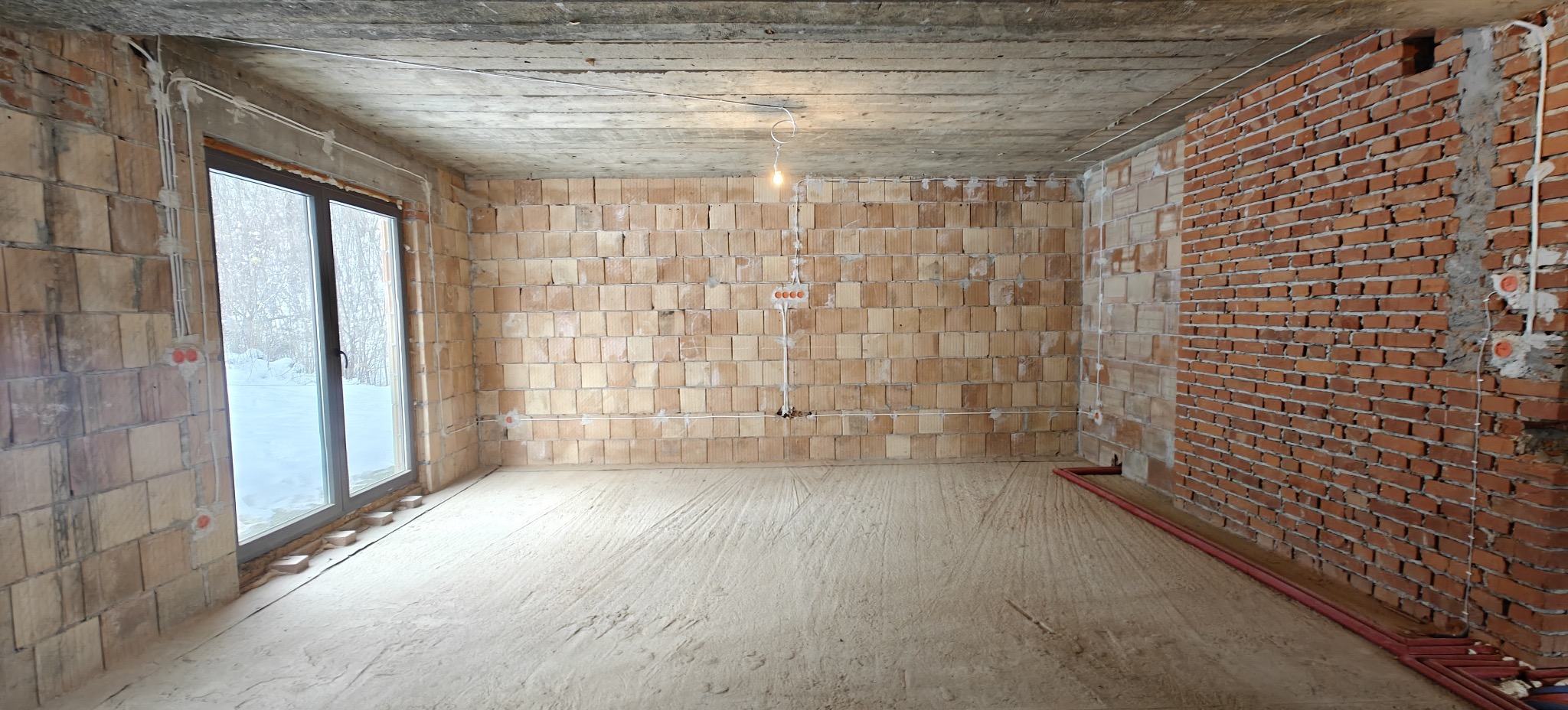Surowy stan wnętrza z widoczną instalacją elektryczną na ścianach z cegły i pustaków, okno z widokiem na zimowy krajobraz, surowy betonowy strop.