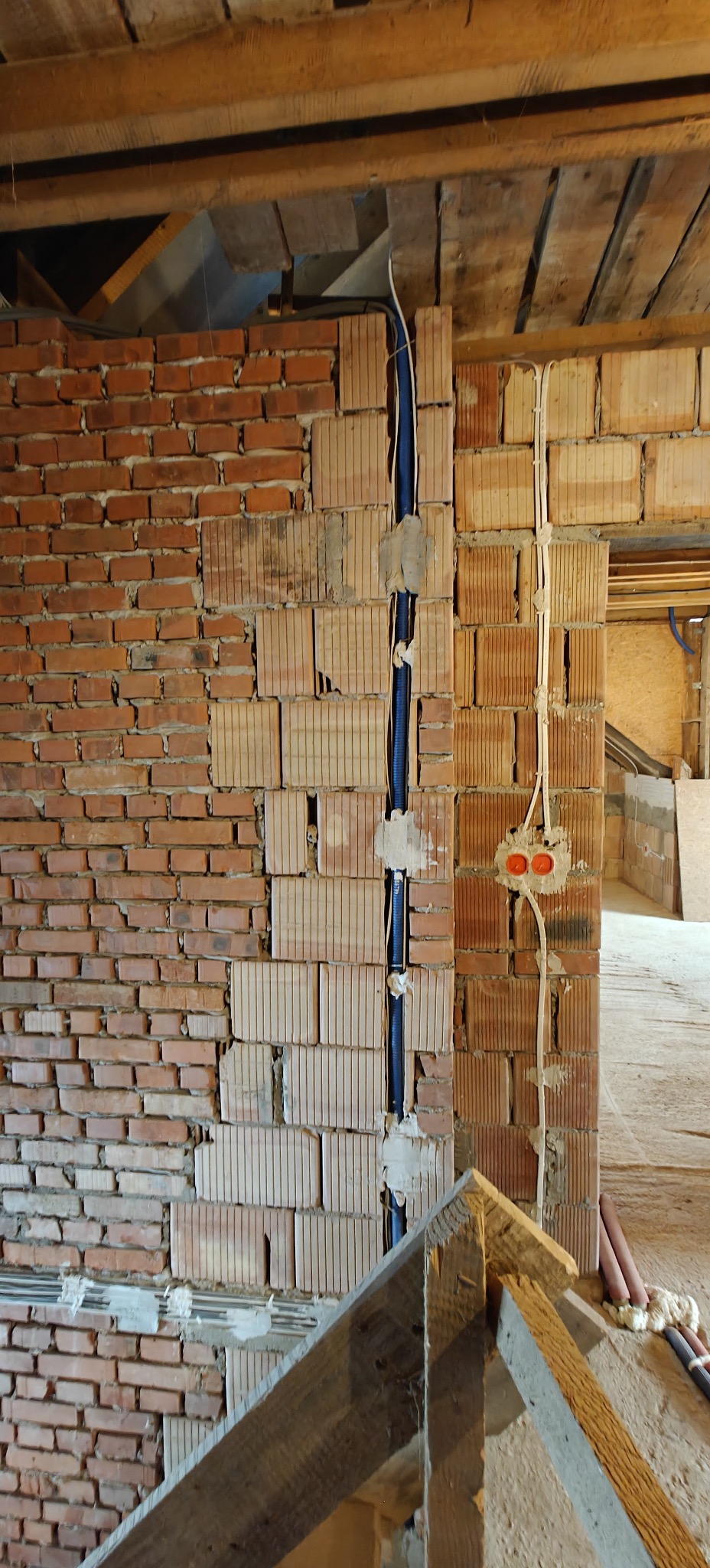 Murowana ściana z widocznymi przewodami w peszlu i puszkami instalacyjnymi. Surowy stan, instalacja w trakcie montażu, widoczne elementy konstrukcji dachu.