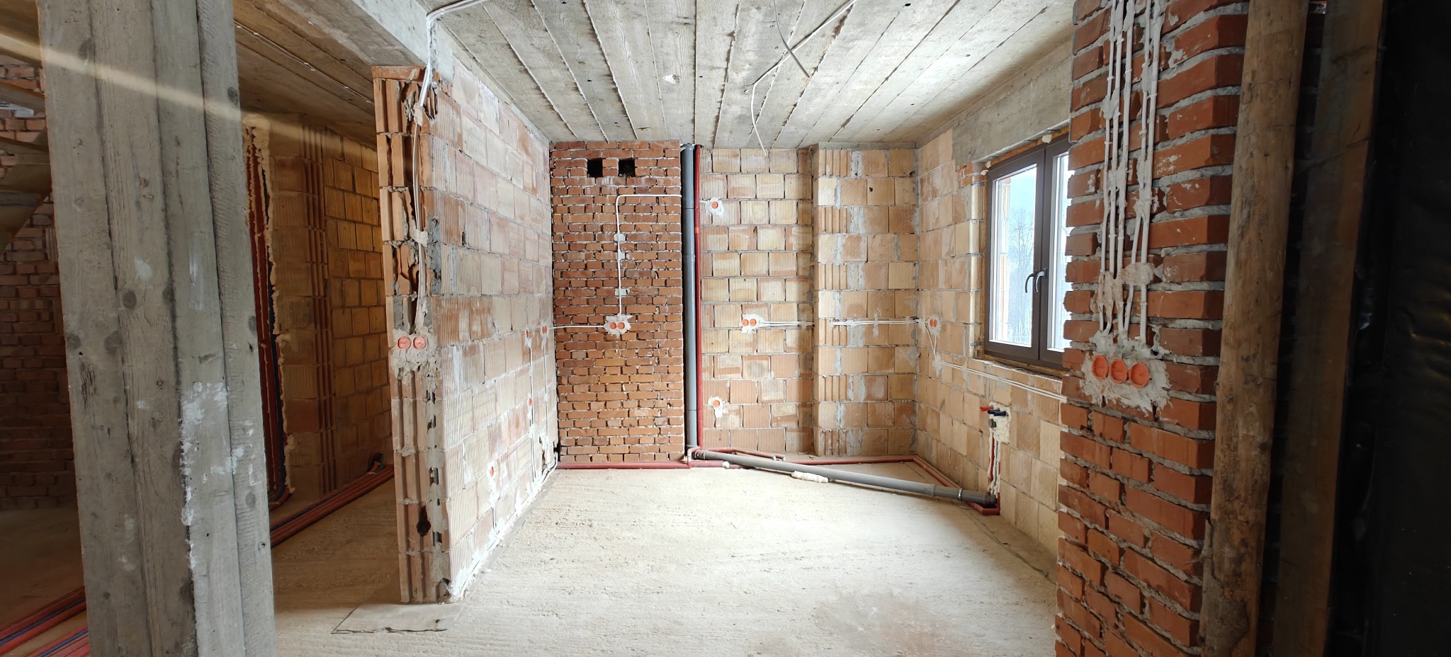 Surowy stan wnętrza z widocznymi instalacjami elektrycznymi w ścianach z cegły dziurawki. Widoczne kable i puszki instalacyjne. W tle okno i surowy betonowy strop.
