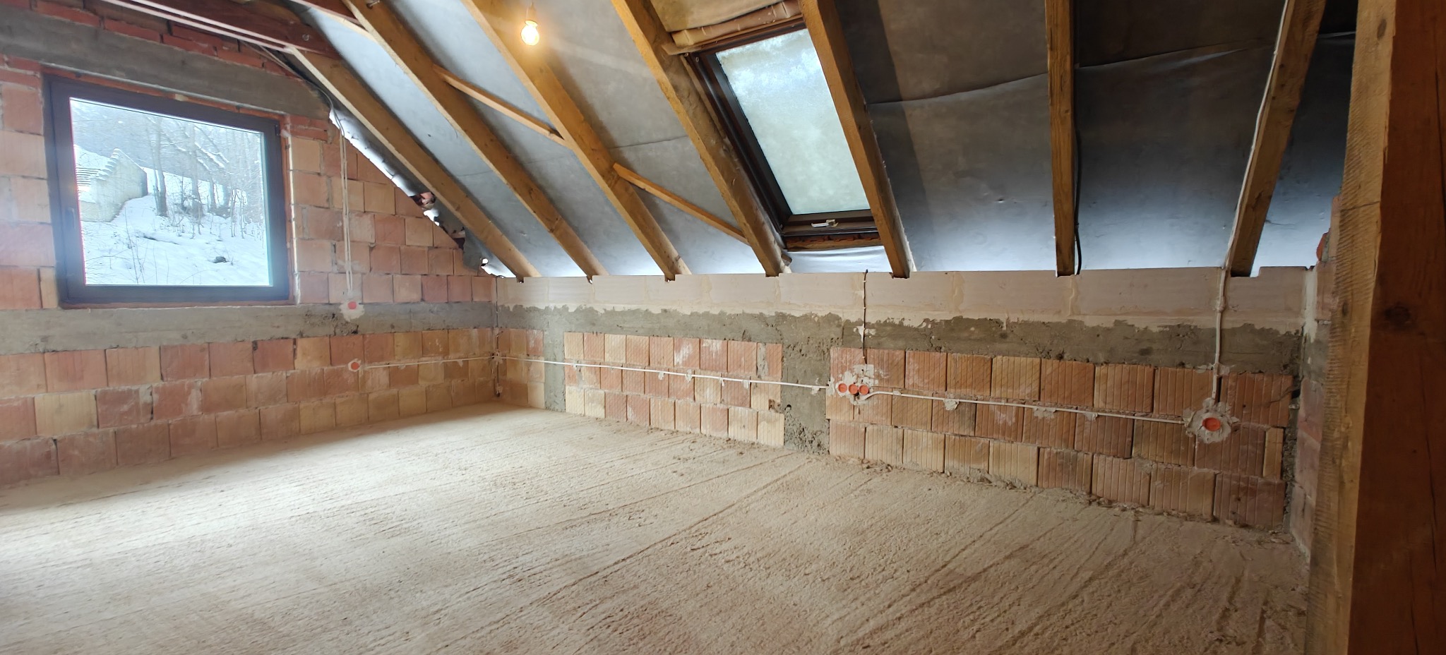 Surowy stan poddasza z oknami i widoczną instalacją elektryczną w postaci przewodów i puszek na ceglanej ścianie. Widoczne drewniane elementy konstrukcji dachu.