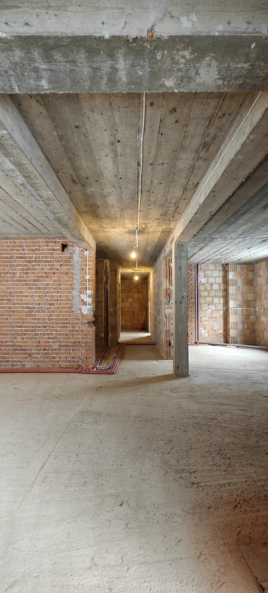 Wnętrze w stanie surowym: widoczne ceglane ściany, betonowy strop z instalacją elektryczną i rurami. W tle perspektywa korytarza z żarówkami.