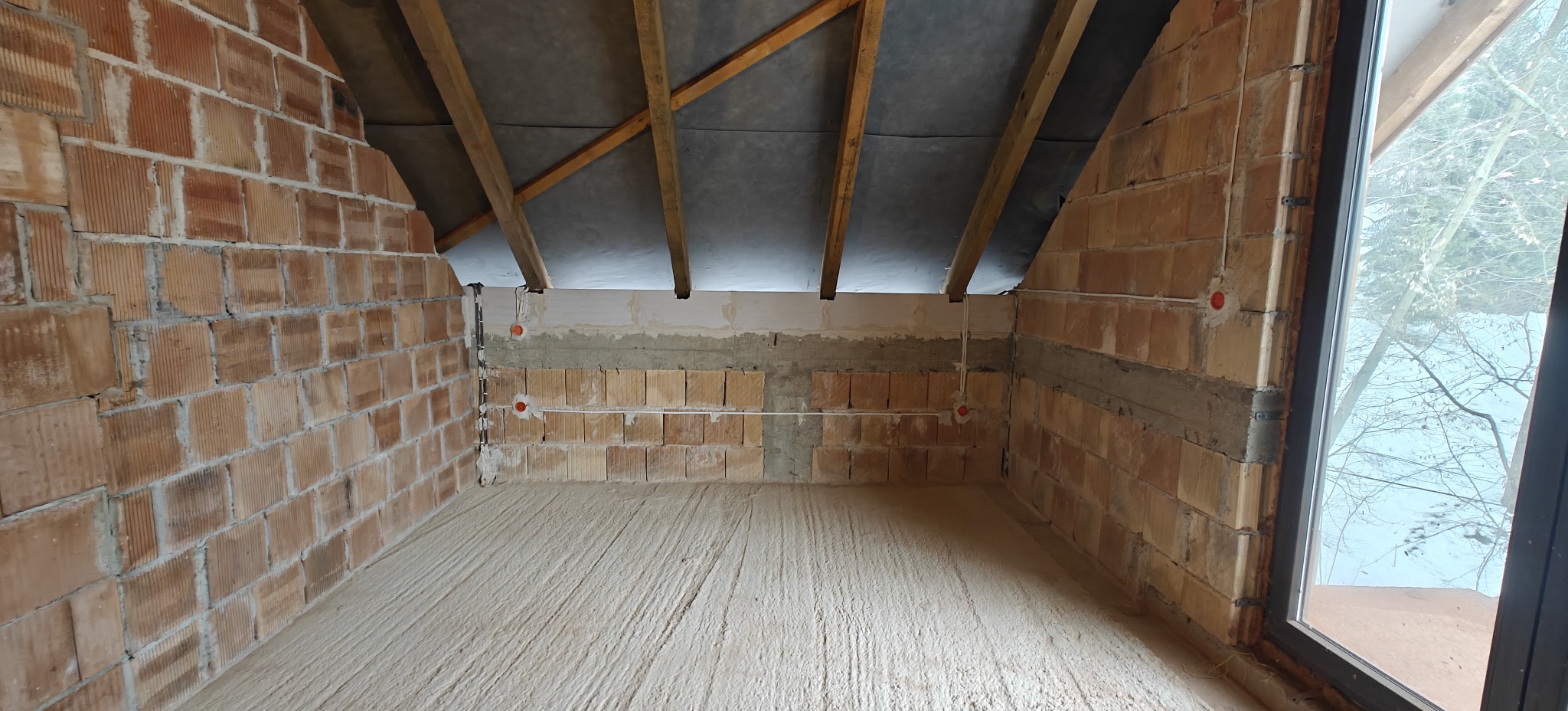 Surowe wnętrze poddasza z ceglanymi ścianami, widocznymi przewodami elektrycznymi i oknem z widokiem na zimowy krajobraz. Wnętrze w trakcie prac wykończeniowych.
