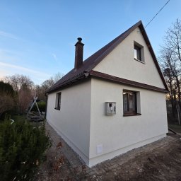 ART-BUD Artur Bomba - Boczny widok domu po renowacji elewacji, z brązowym dachem i oknami. Świeżo otynkowana, jasna ściana kontrastuje z otoczeniem ogrodu i błękitnym niebem.