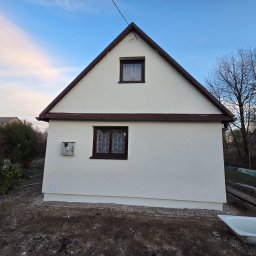 ART-BUD Artur Bomba - Biały dom z brązowym dachem po renowacji elewacji, widoczne dwa okna i skrzynka elektryczna. Tło: błękitne niebo i drzewa bez liści. Ujęcie z dołu.