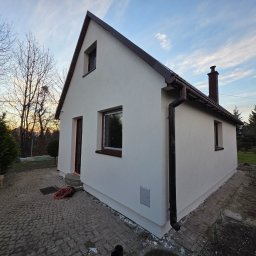 ART-BUD Artur Bomba - Otynkowany, parterowy dom z brązowym dachem i rynnami, sfotografowany z niskiego kąta. Widoczne okna z brązowymi ramami i komin. Na pierwszym planie kostka brukowa.