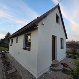 ART-BUD Artur Bomba - Otynkowany dom jednorodzinny z brązowym dachem i oknami, widok z boku na tle błękitnego nieba o zmierzchu. Widoczny chodnik z kostki i elementy ogrodu.