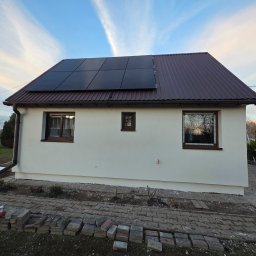 ART-BUD Artur Bomba - Biały dom z brązowym dachem i panelami słonecznymi. Na pierwszym planie kostka brukowa i cegły. Widoczne okna z brązowymi ramami i rynna.