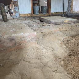 ART-BUD Artur Bomba - Przygotowanie podłoża pod posadzkę w Biłgoraju: wykopana ziemia, odsłonięte cegły fundamentów, widoczne ślady stóp. Dwa wiadra i poziomica w tle.