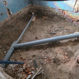 ART-BUD Artur Bomba - Szare rury kanalizacyjne w wykopie, fragment niebieskich płytek na ścianie, gruz i narzędzia. Widoczne tęczowe refleksy światła na ścianie.