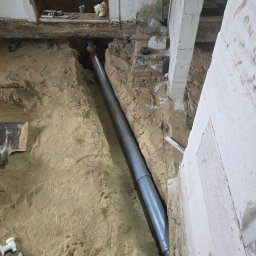 ART-BUD Artur Bomba - Szara rura kanalizacyjna ułożona w wykopie z piasku wewnątrz budynku. Widoczne elementy instalacyjne i narzędzia. Prace instalacyjne w trakcie remontu.