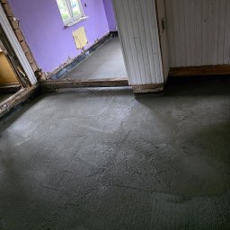 ART-BUD Artur Bomba - Wylany, świeży beton na podłodze w remontowanym pomieszczeniu z fioletowymi ścianami i drewnianymi elementami konstrukcyjnymi. Widoczne ślady po rozprowadzaniu betonu.