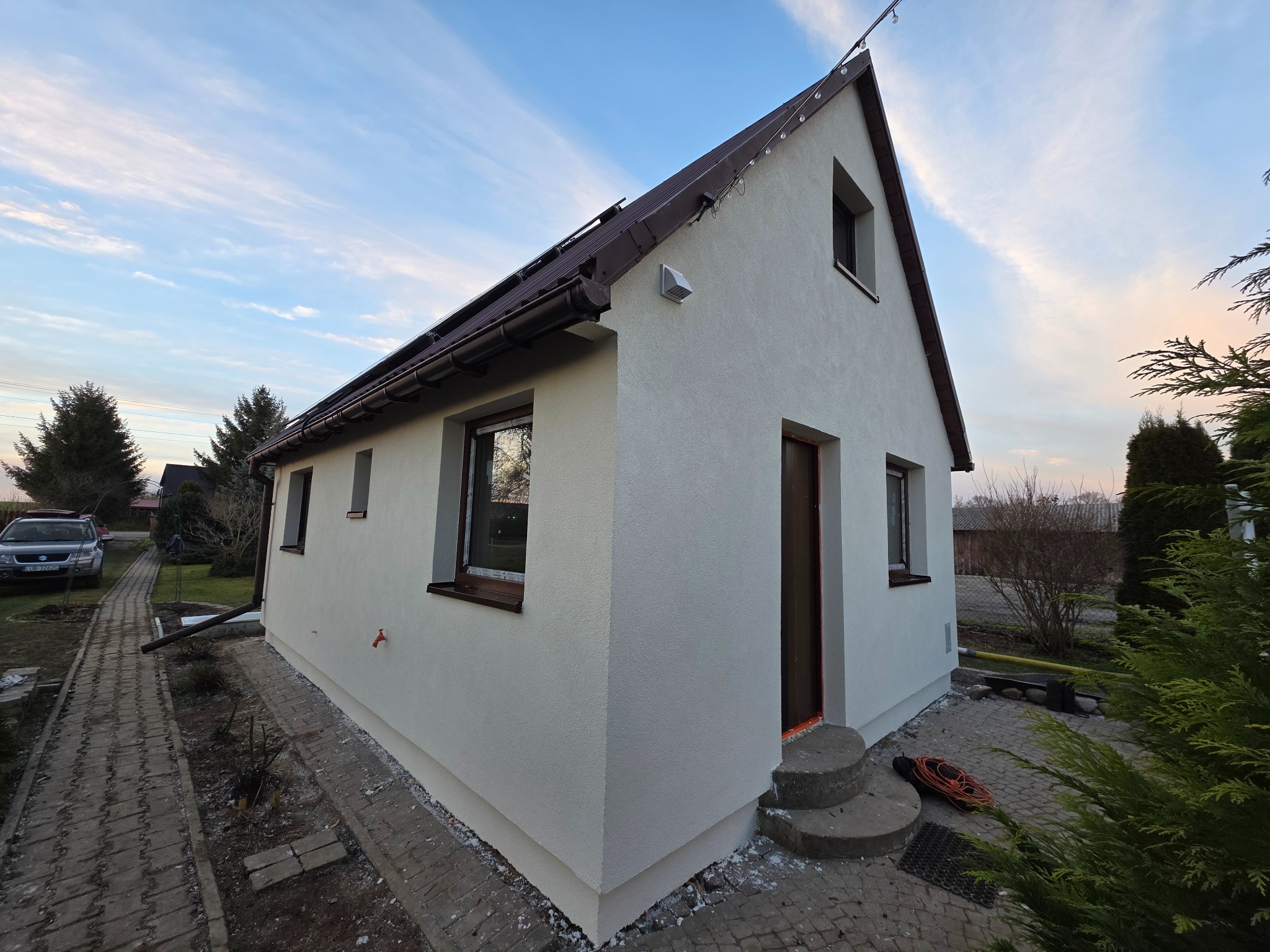 Dom jednorodzinny po renowacji elewacji, z nowym tynkiem i pomalowanymi oknami, widoczny z perspektywy ogrodu w słoneczny dzień. Detal: brązowe rynny.