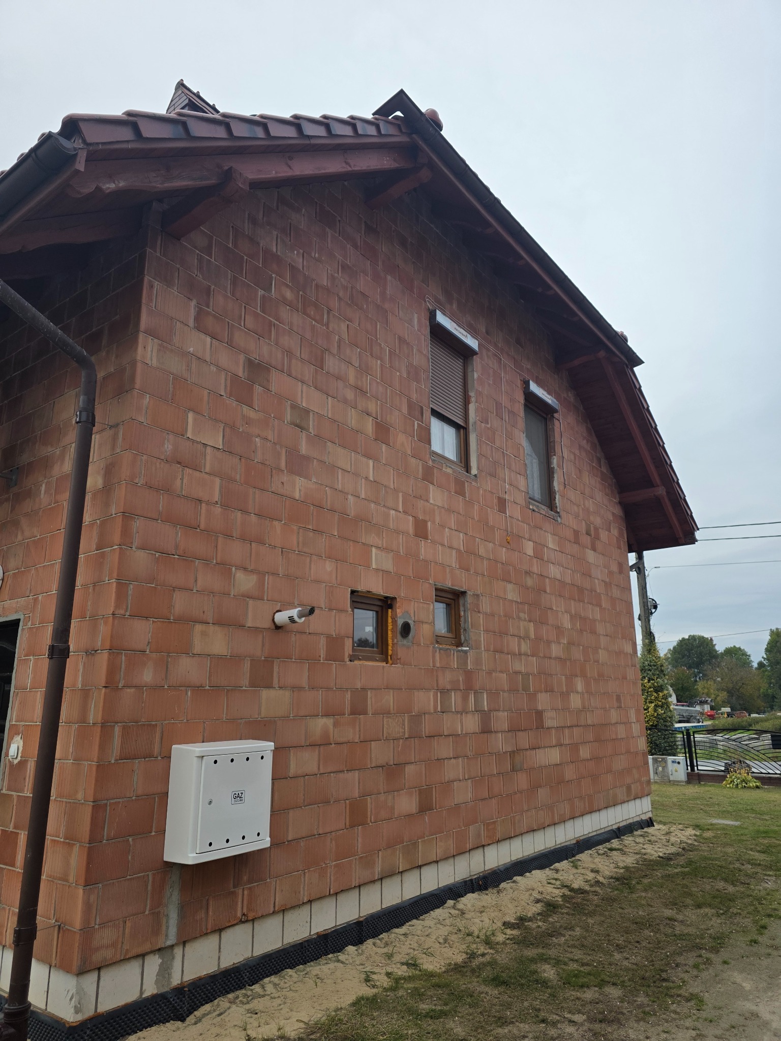 Surowa elewacja z cegły ceramicznej domu jednorodzinnego z zamontowaną skrzynką gazową i widocznymi otworami wentylacyjnymi. Budynek w trakcie budowy.