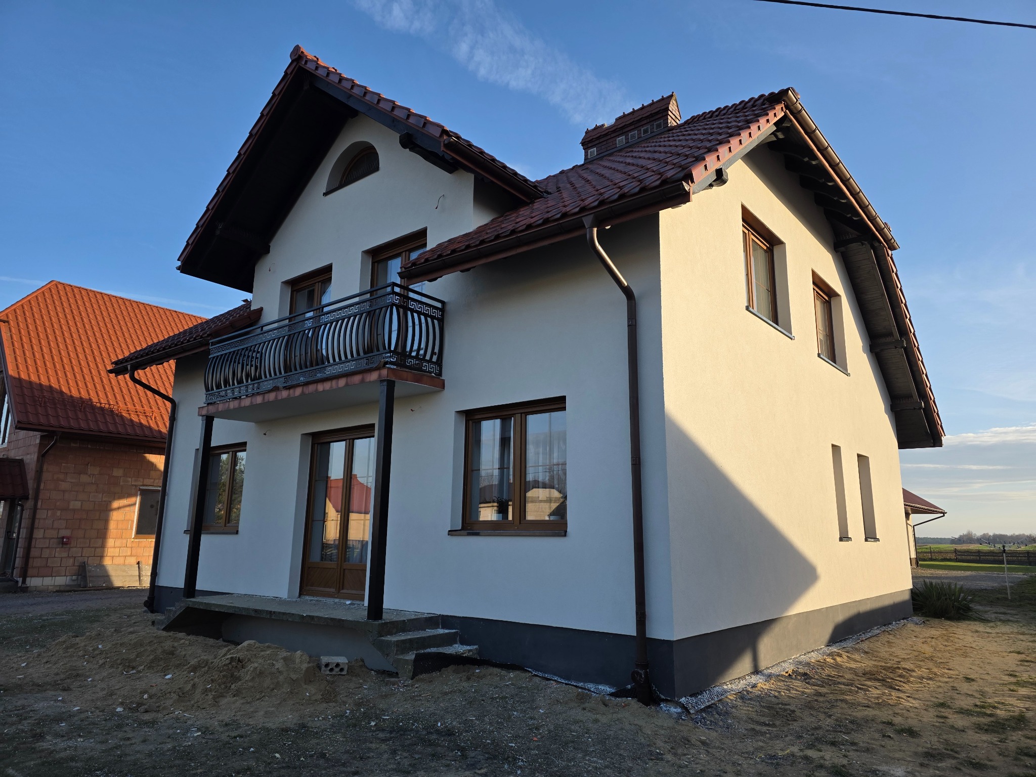 Dwupiętrowy dom z balkonem i brązowym dachem, świeżo po pracach elewacyjnych. Jasna fasada w kontraście z ciemną podmurówką i błękitnym niebem.