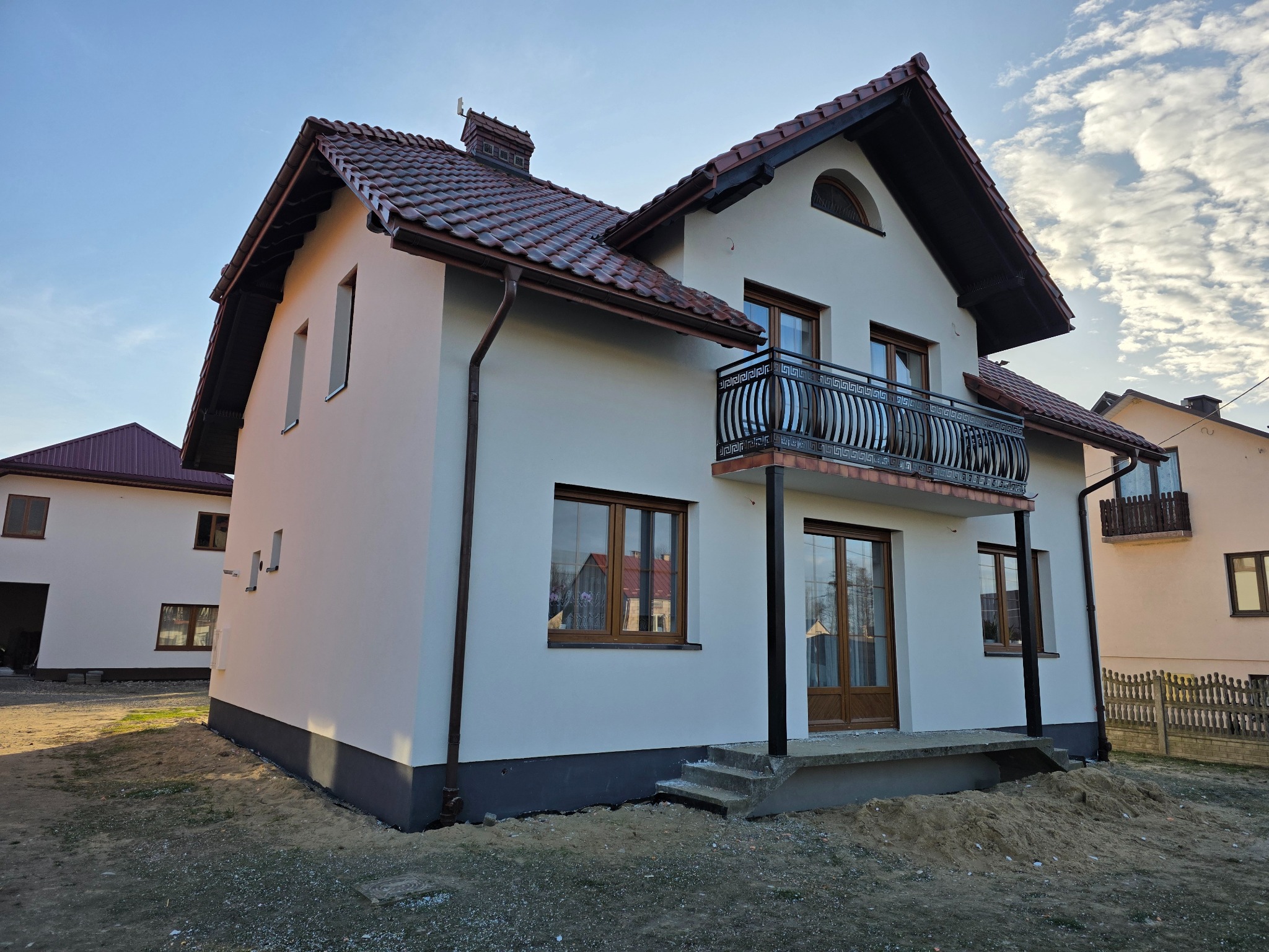Biały dom jednorodzinny z balkonem i brązowym dachem, po renowacji elewacji. Widoczne okna z brązowymi ramami i fragment niezagospodarowanego terenu.