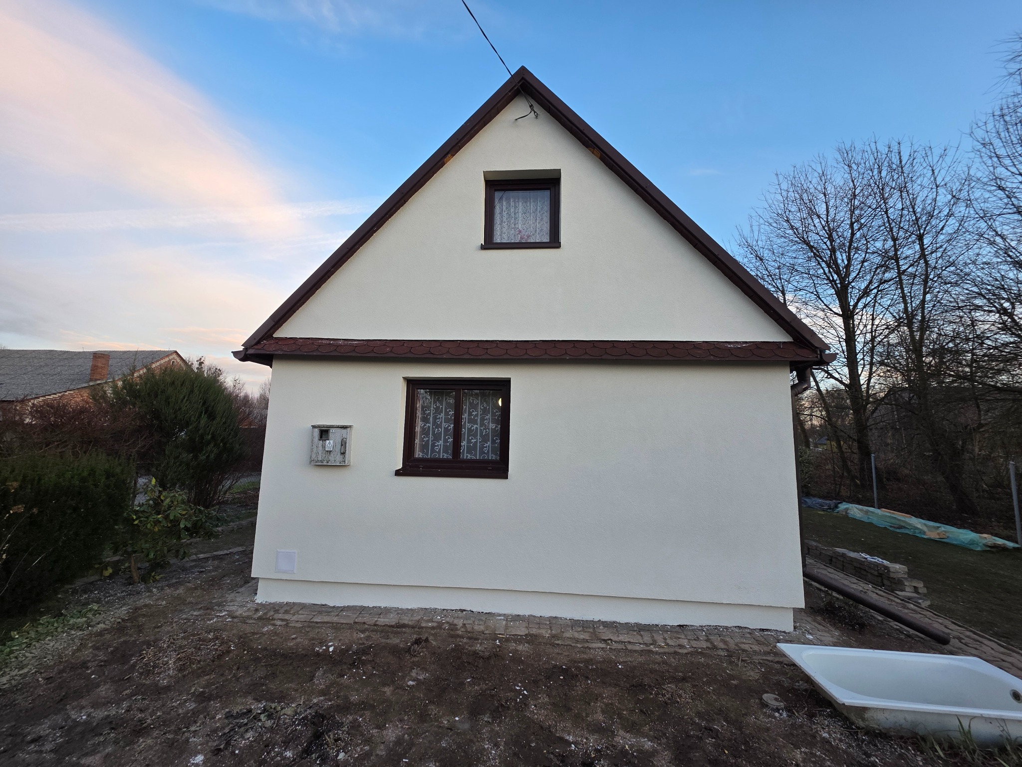 Biały dom z brązowym dachem po renowacji elewacji, widoczne dwa okna i skrzynka elektryczna. Tło: błękitne niebo i drzewa bez liści. Ujęcie z dołu.