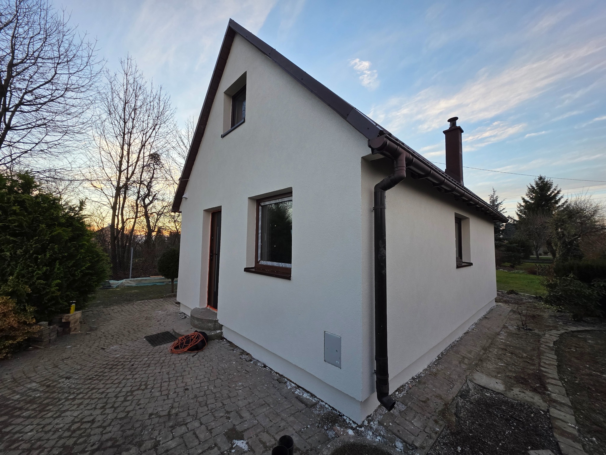 Otynkowany, parterowy dom z brązowym dachem i rynnami, sfotografowany z niskiego kąta. Widoczne okna z brązowymi ramami i komin. Na pierwszym planie kostka brukowa.