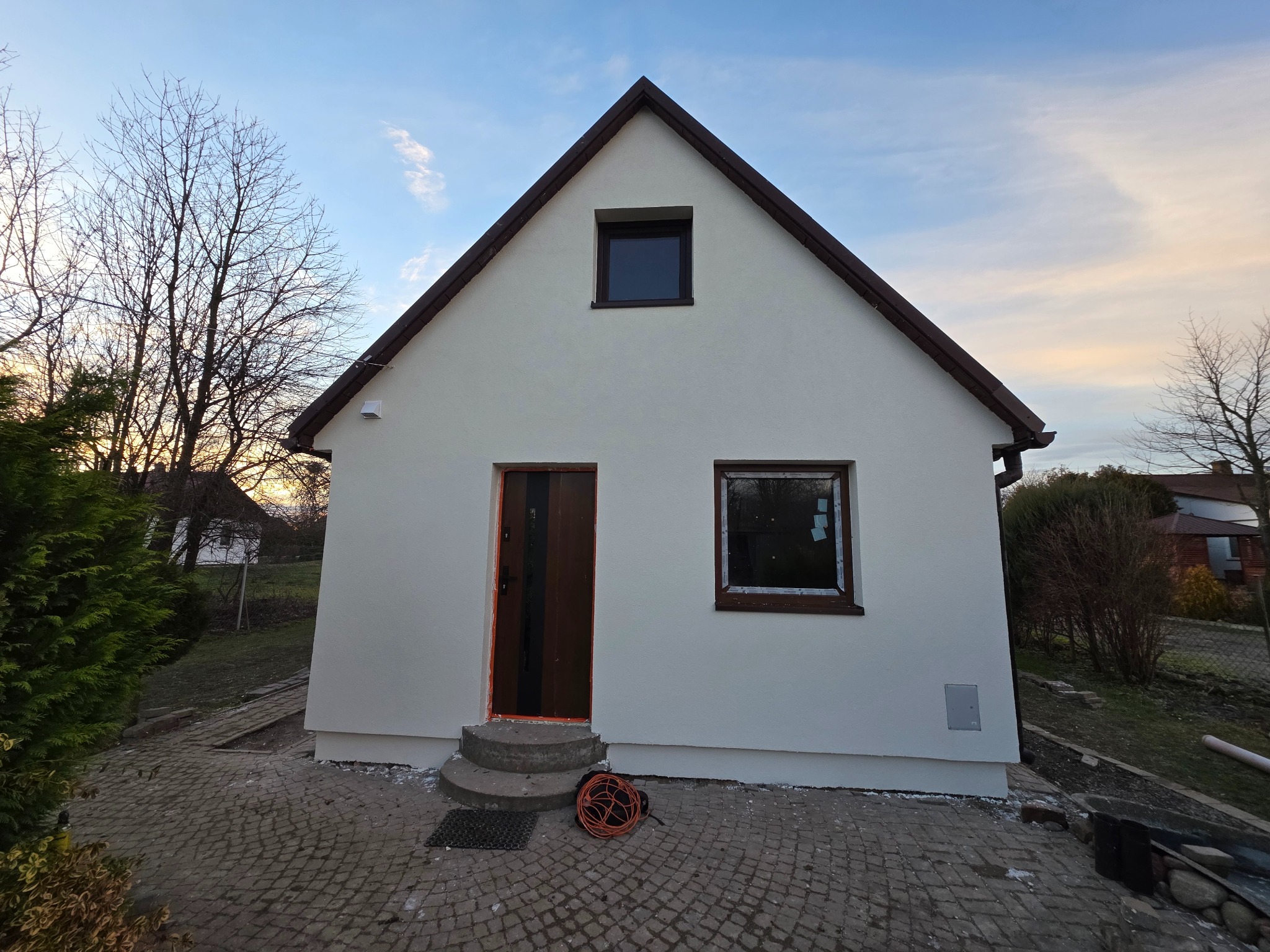 Biały dom jednorodzinny po tynkowaniu elewacji, z brązowymi ramami okiennymi i drzwiami. Widoczne schody wejściowe i kostka brukowa. Częściowo zachmurzone niebo.