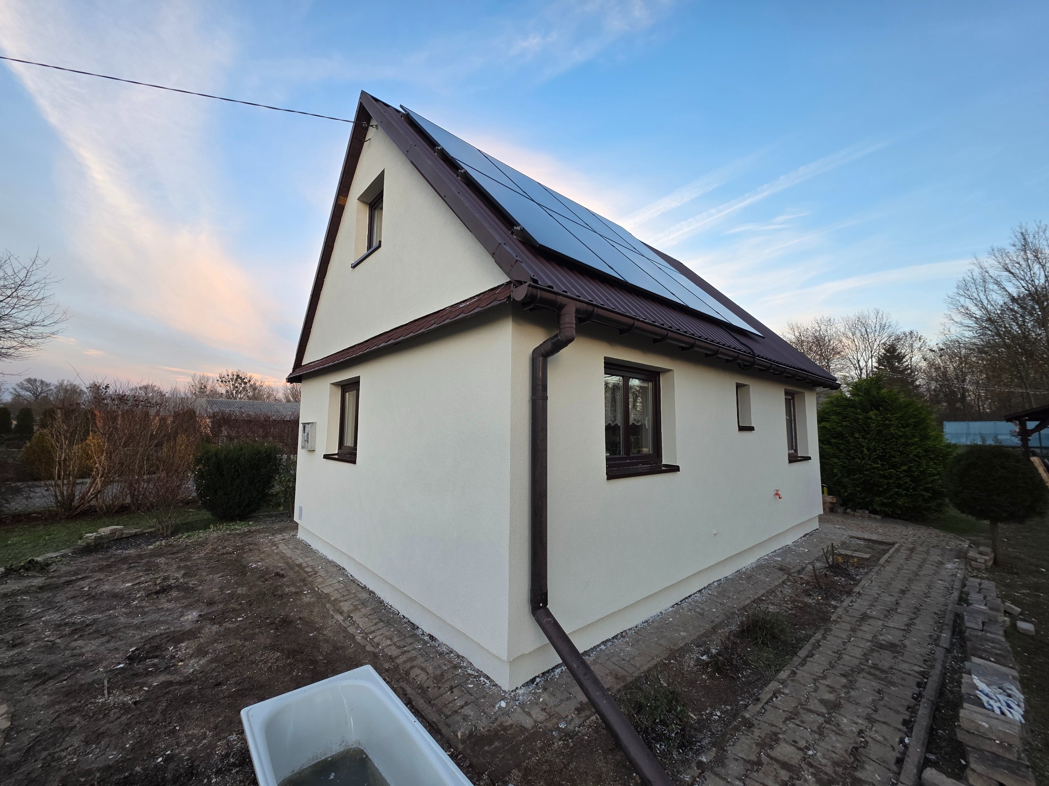 Dom jednorodzinny po renowacji elewacji, z nowymi panelami słonecznymi na dachu. Widoczne świeże tynki i brązowe rynny. Ujęcie z poziomu gruntu, błękitne niebo w tle.