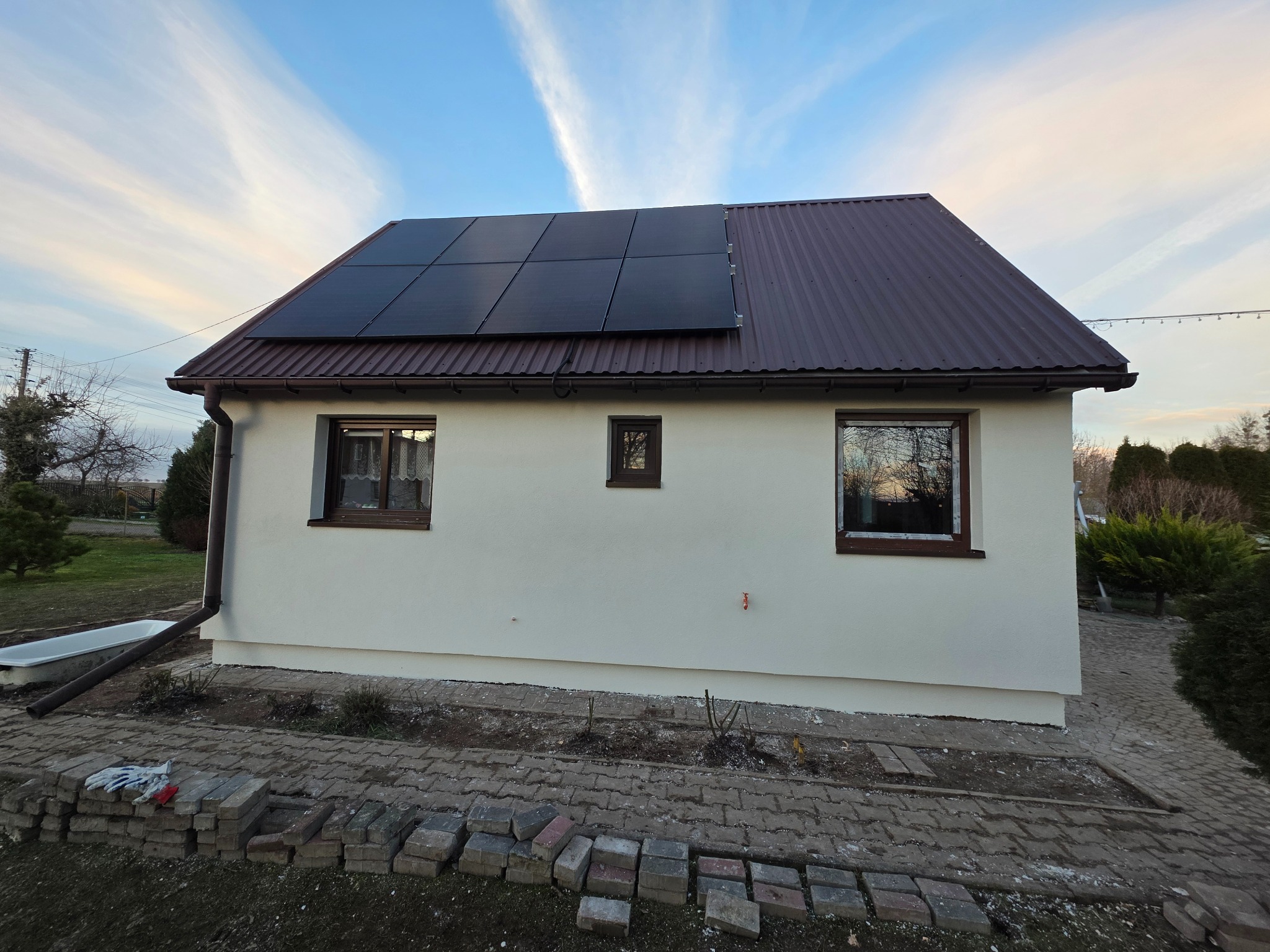 Biały dom z brązowym dachem i panelami słonecznymi. Na pierwszym planie kostka brukowa i cegły. Widoczne okna z brązowymi ramami i rynna.