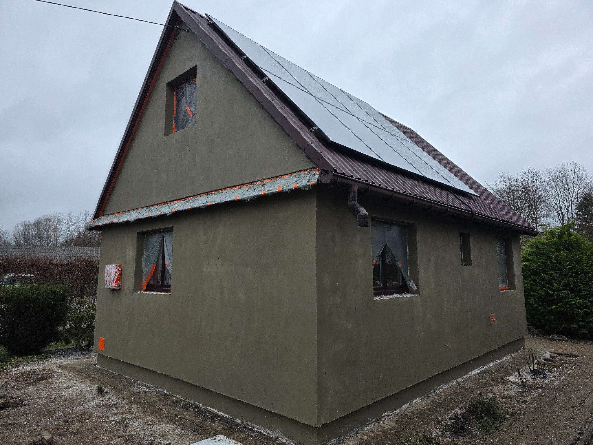 Niski dom z szarą elewacją w trakcie prac wykończeniowych, okna zabezpieczone folią, panele słoneczne na dachu. Widok z zewnątrz, pochmurny dzień.
