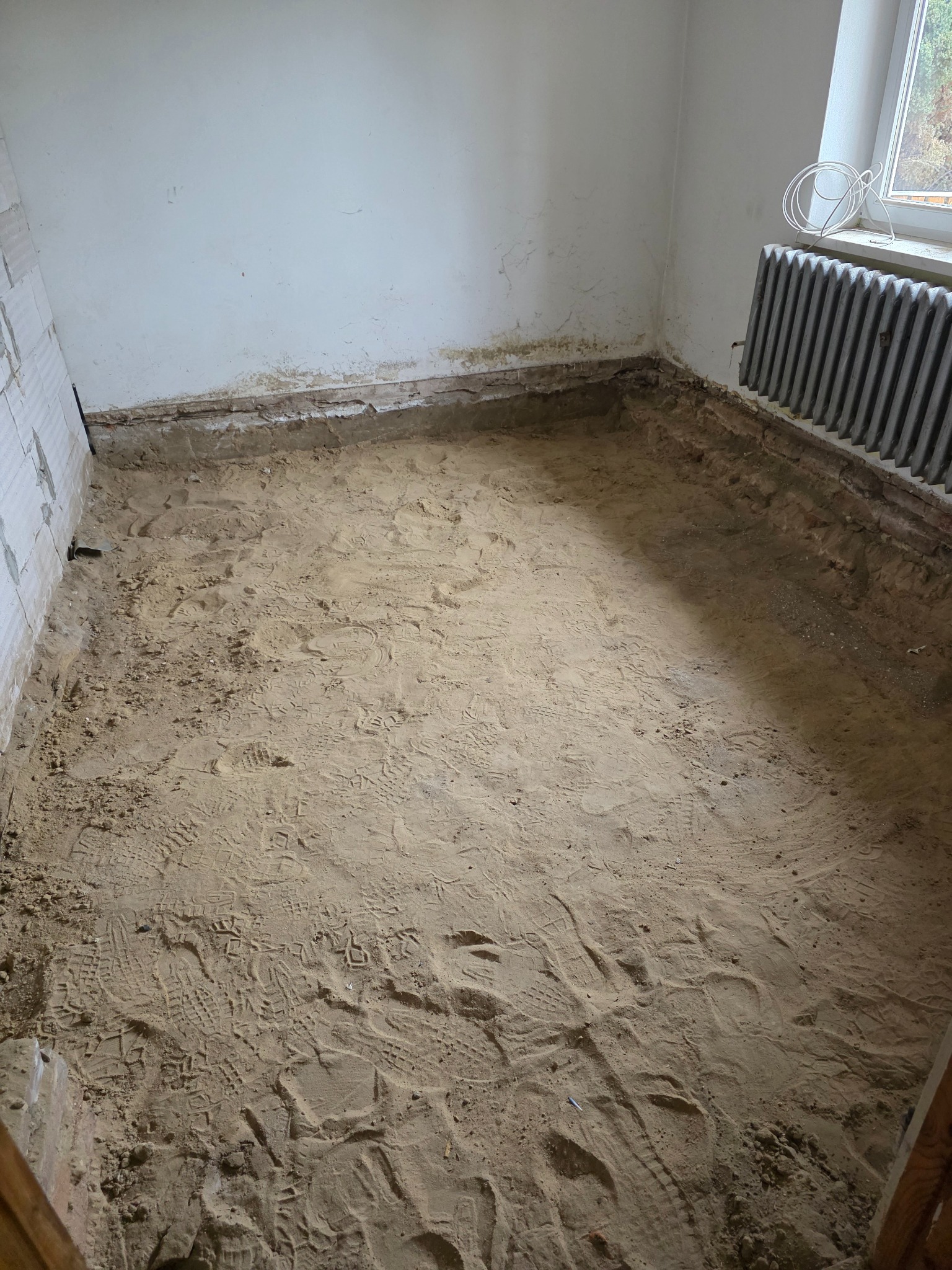Przygotowanie podłoża pod wylewkę w pomieszczeniu z grzejnikiem. Widoczne ślady butów na piasku, fragment okna i ściany z resztkami starej farby.