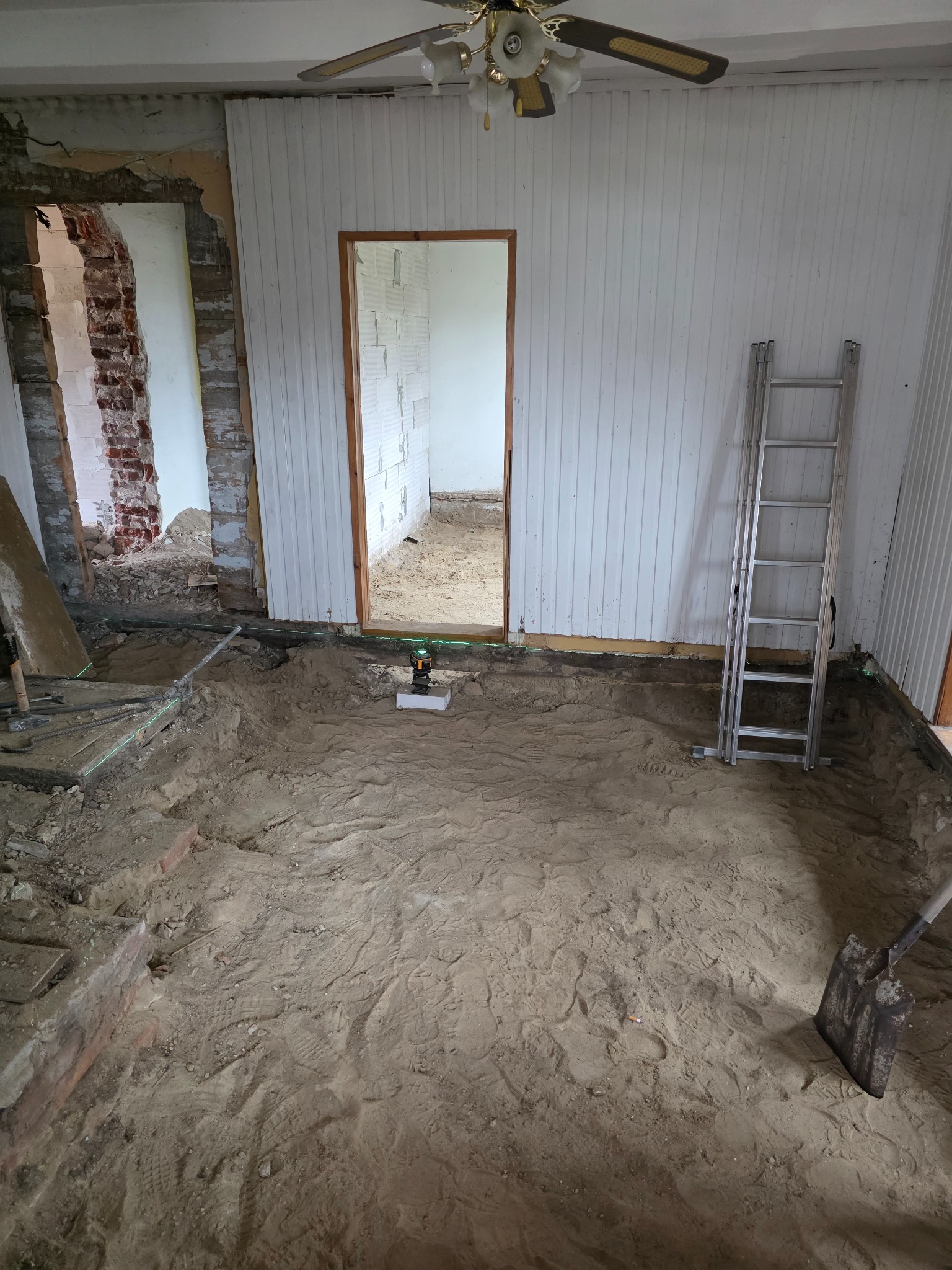 Wnętrze w trakcie remontu: odsłonięte cegły, ślady po usuniętej podłodze, laser krzyżowy, drabina oparta o ścianę z boazerią. Widoczne narzędzia i ślady stóp w piasku.