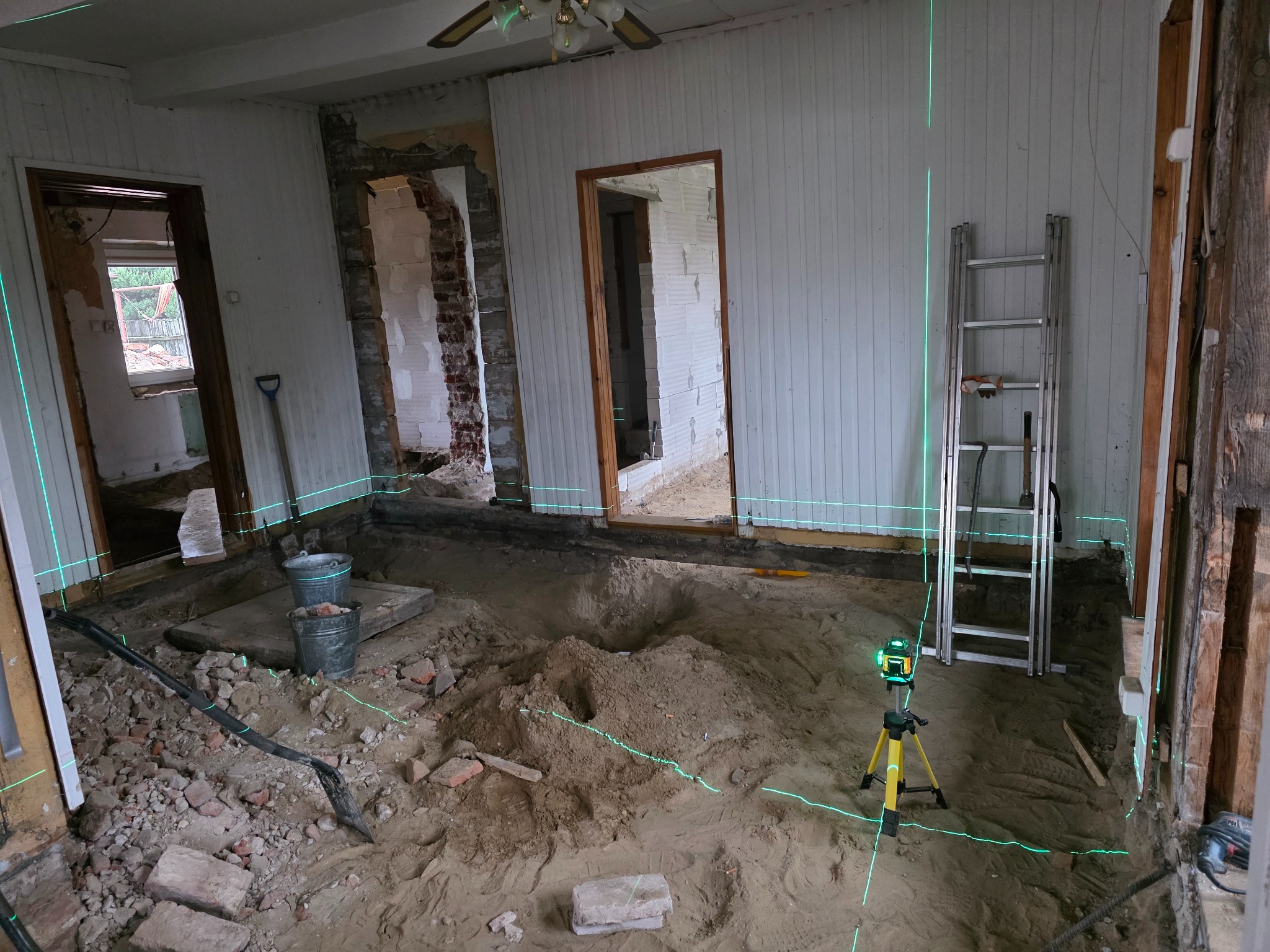 Wnętrze w trakcie remontu: wykopana podłoga, cegły, poziomica laserowa, drabina. Widoczne ślady demontażu ścian i podłóg. Przygotowanie do dalszych prac.