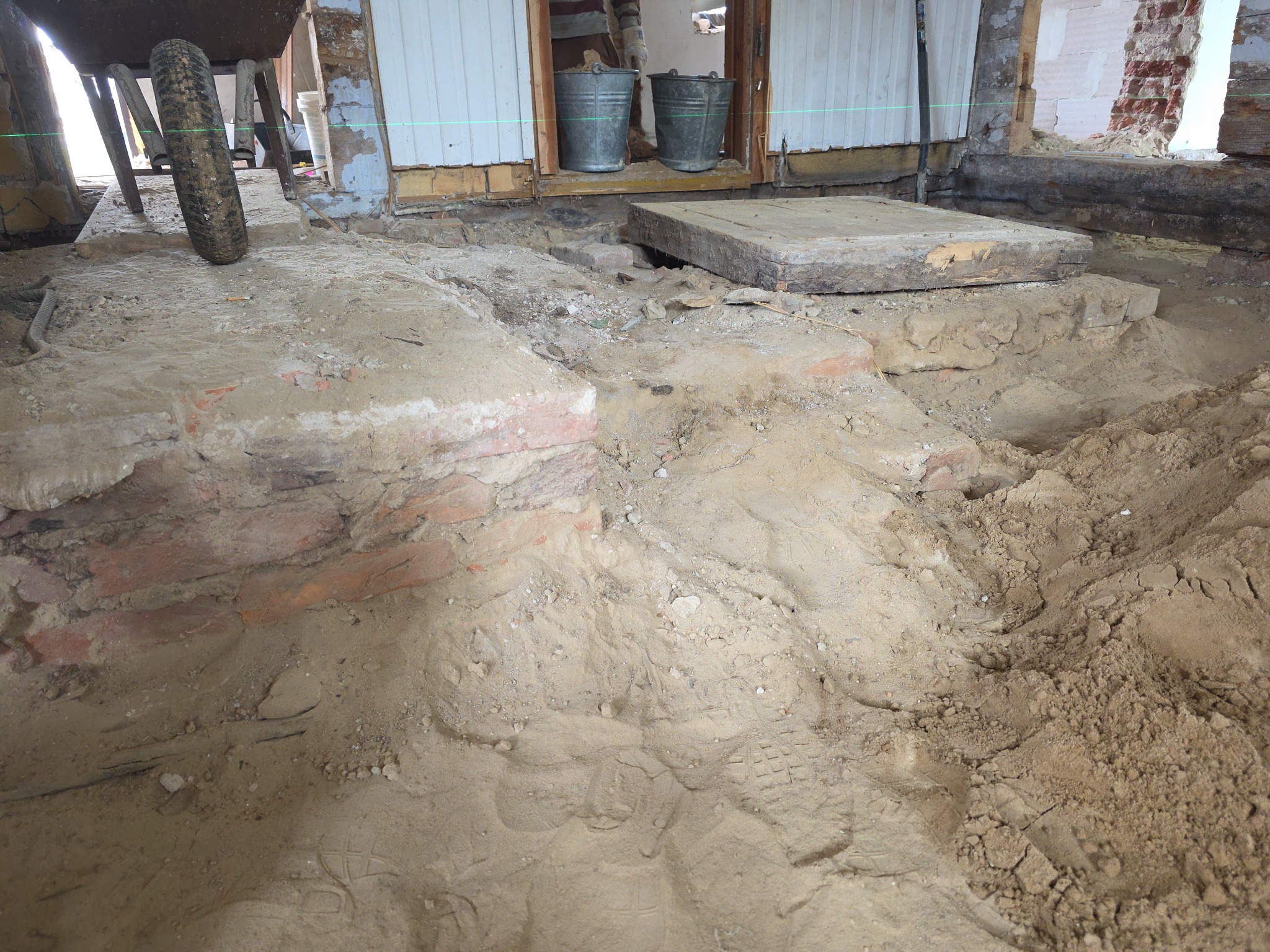 Przygotowanie podłoża pod posadzkę w Biłgoraju: wykopana ziemia, odsłonięte cegły fundamentów, widoczne ślady stóp. Dwa wiadra i poziomica w tle.