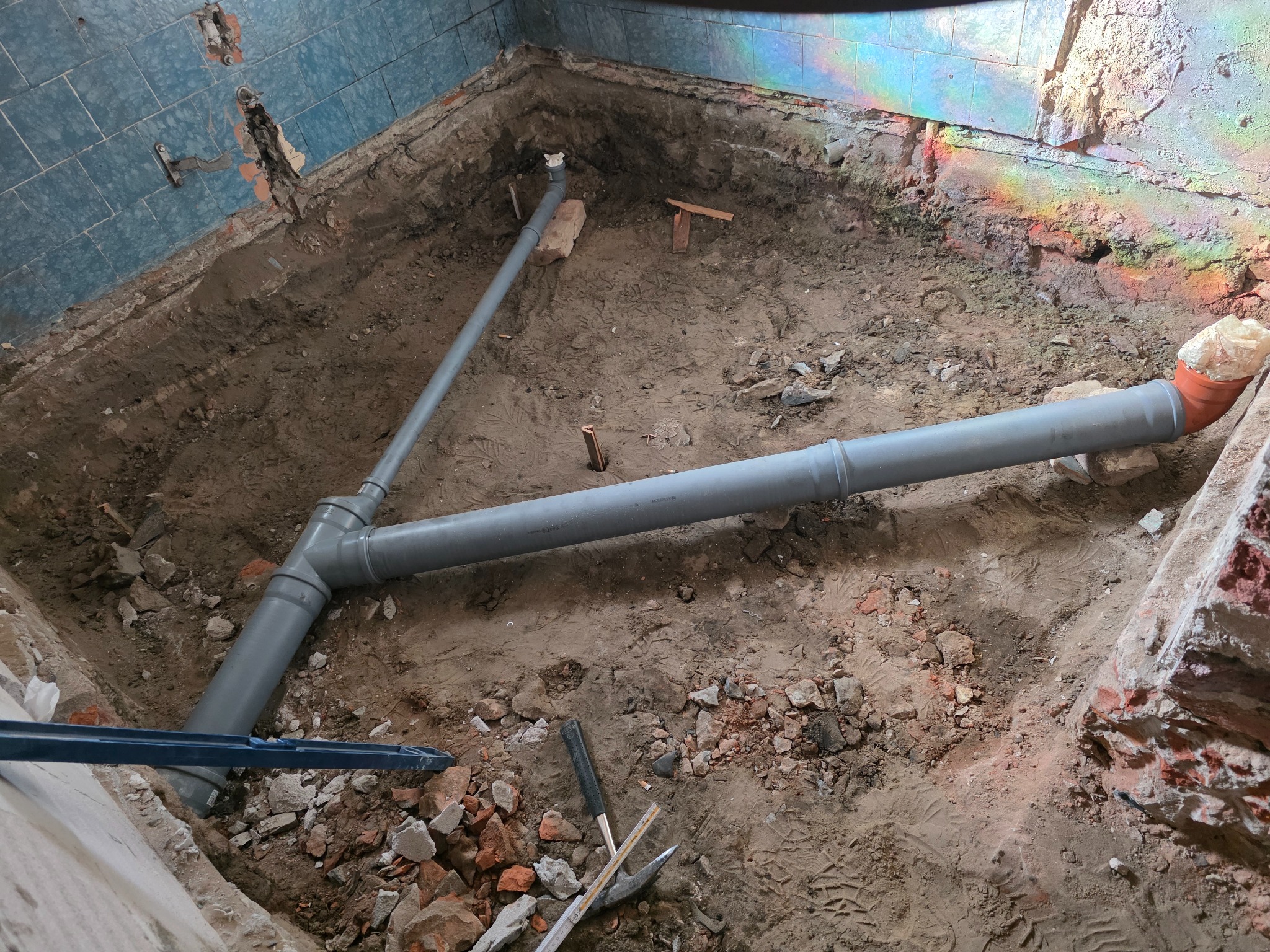 Szare rury kanalizacyjne w wykopie, fragment niebieskich płytek na ścianie, gruz i narzędzia. Widoczne tęczowe refleksy światła na ścianie.