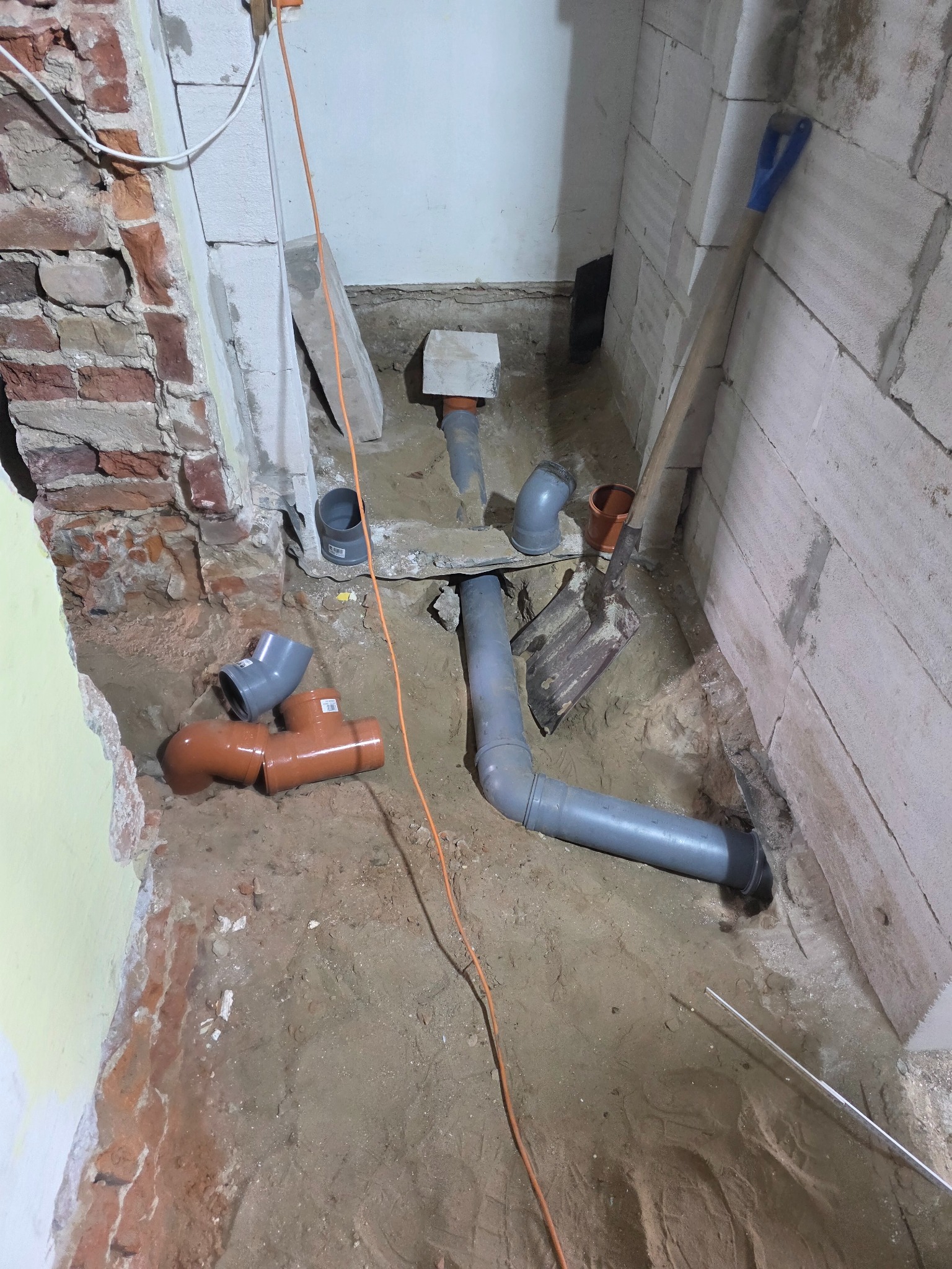 Instalacja kanalizacyjna w trakcie remontu łazienki: szare i brązowe rury PVC ułożone w wykopie, widoczna cegła i bloczki, łopata oparta o ścianę.