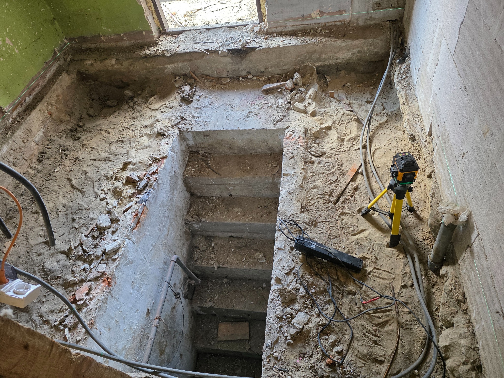 Wnętrze w trakcie remontu: wykopana dziura w podłodze, widoczne przewody, rury i poziomica laserowa na statywie. Surowy stan, przygotowanie do dalszych prac.