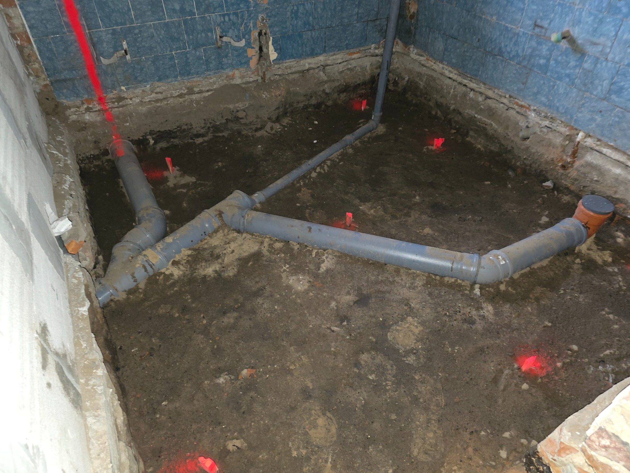 Szare rury kanalizacyjne ułożone w wykopie, z widocznymi czerwonymi znacznikami laserowymi, na tle niebieskich, uszkodzonych płytek w trakcie remontu łazienki.