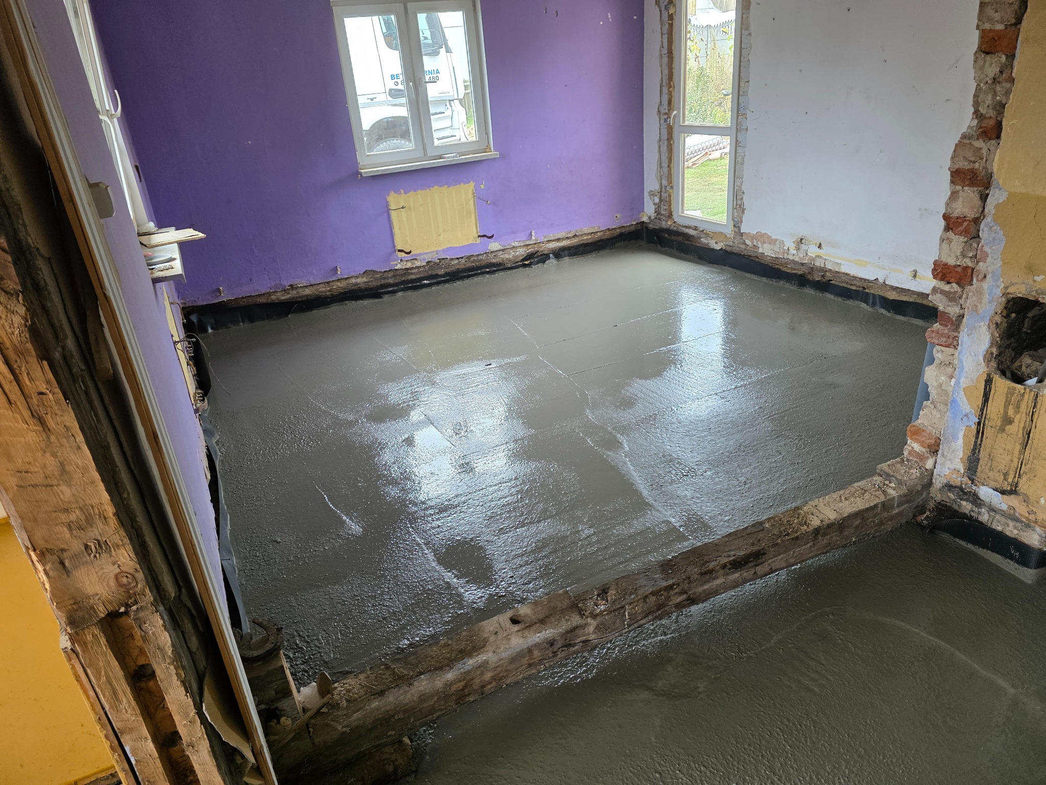 Wylany beton na podłodze w trakcie remontu mieszkania. Widoczne fragmenty ścian w kolorze fioletowym i białym, okna oraz drewniane elementy konstrukcyjne.