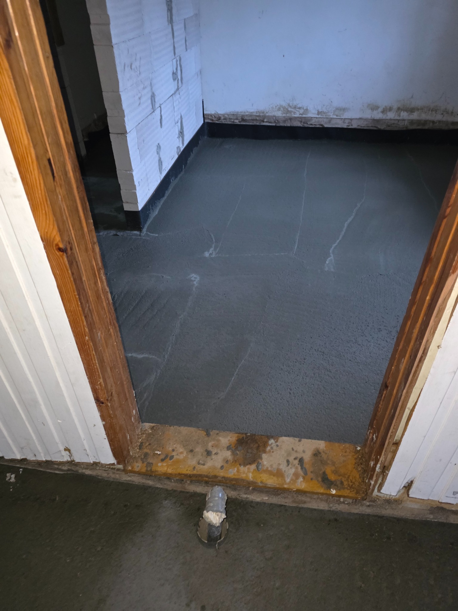 Wylana, świeża wylewka betonowa w pomieszczeniu z widocznymi bloczkami z betonu komórkowego i drewnianą futryną drzwiową. Widoczne ślady po łatach.