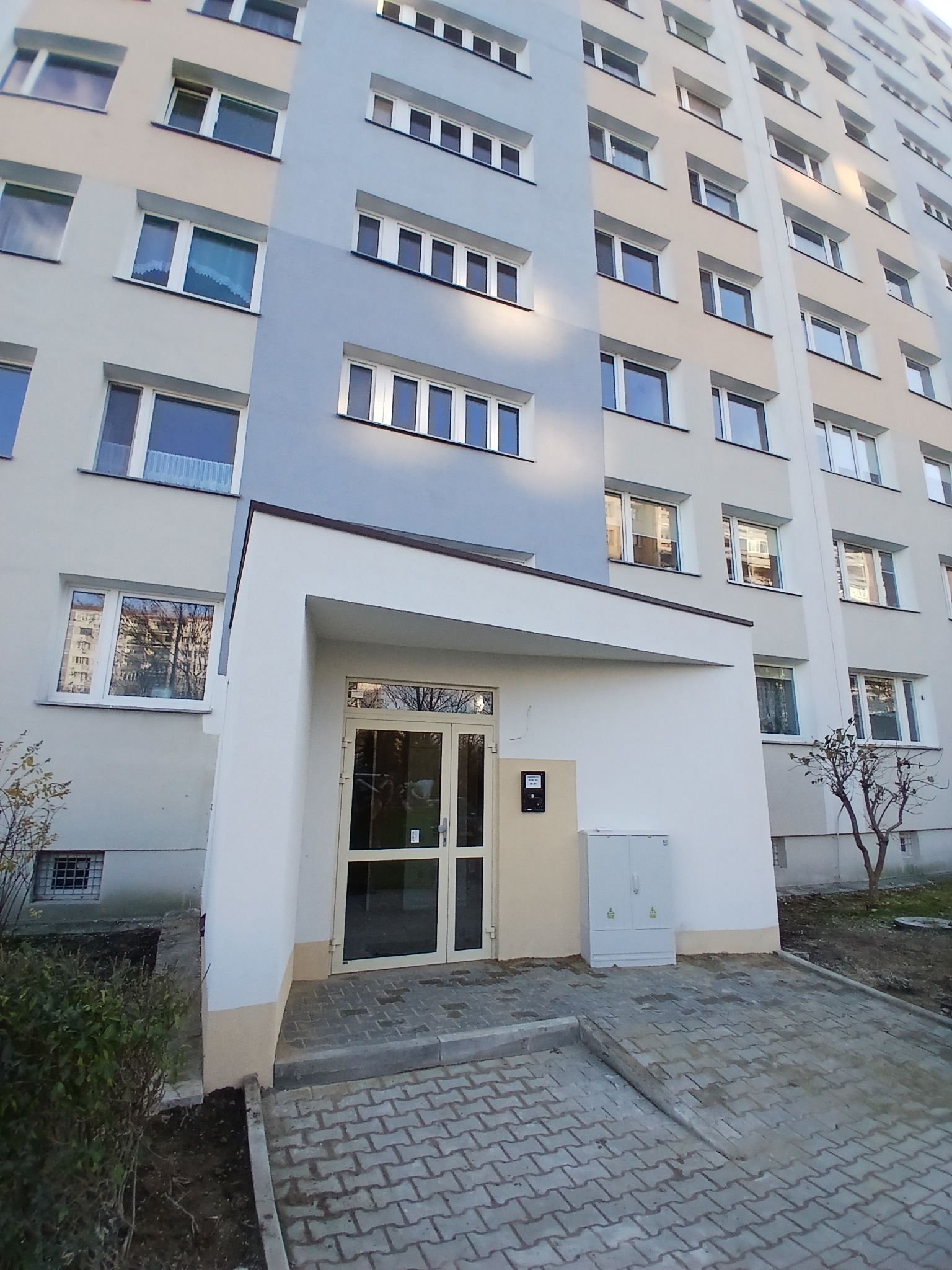 Odnowiona elewacja bloku z wejściem, świeżo pomalowana na szaro i biało, z nową kostką brukową przed wejściem. Widoczne okna i drzwi wejściowe.