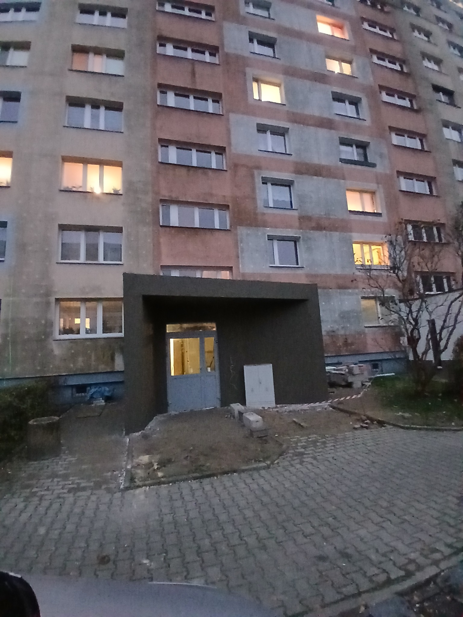 Odnowione wejście do bloku z nową, ciemnoszarą konstrukcją nad drzwiami. Widoczne materiały budowlane i kostka brukowa. W tle oświetlony budynek mieszkalny.