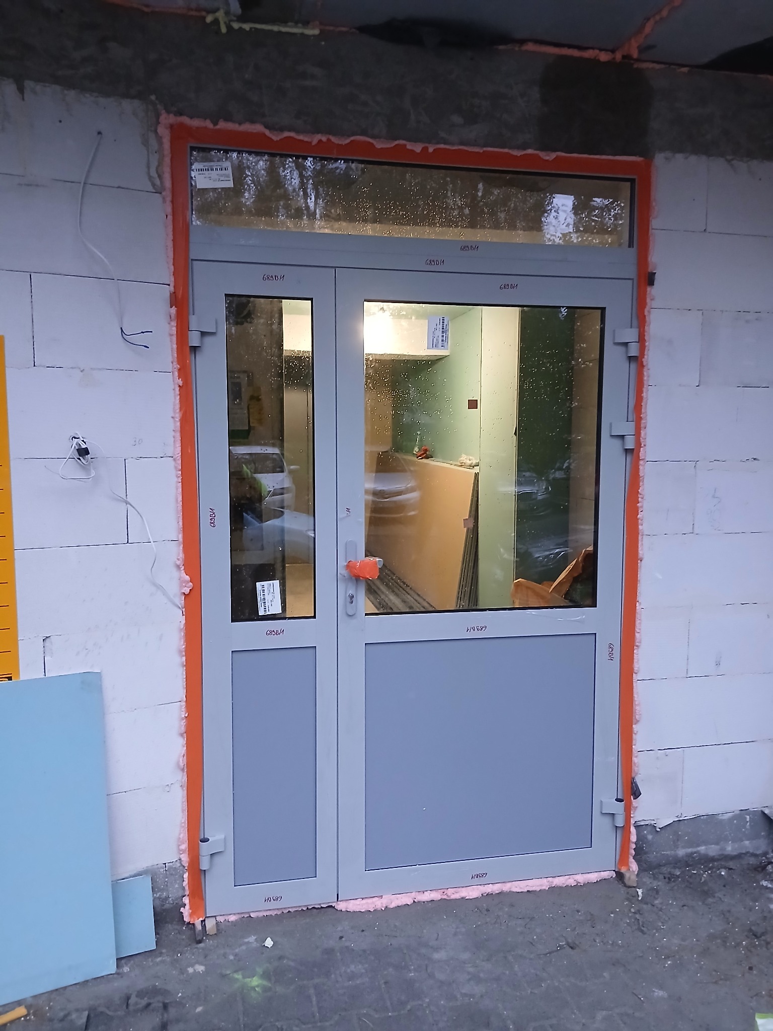 Szare, dwuskrzydłowe drzwi wejściowe z przeszkleniami, osadzone w surowym murze z białych bloczków, uszczelnione pianką montażową. Widoczne płyty karton-gips wewnątrz.