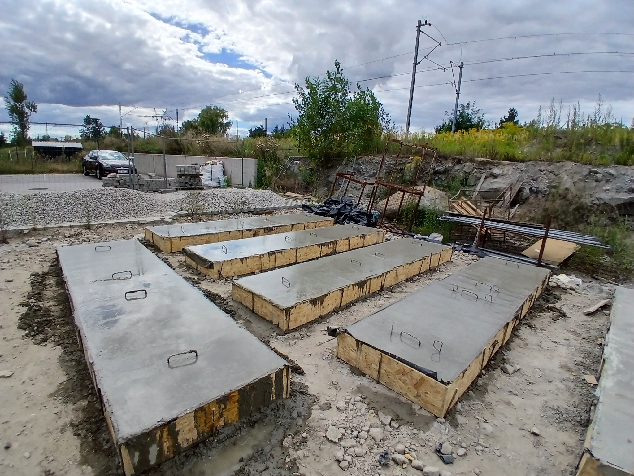 Świeżo wylane, prostokątne fundamenty w drewnianych szalunkach, z metalowymi uchwytami, na placu budowy. W tle materiały budowlane i krajobraz z liniami wysokiego napięcia.