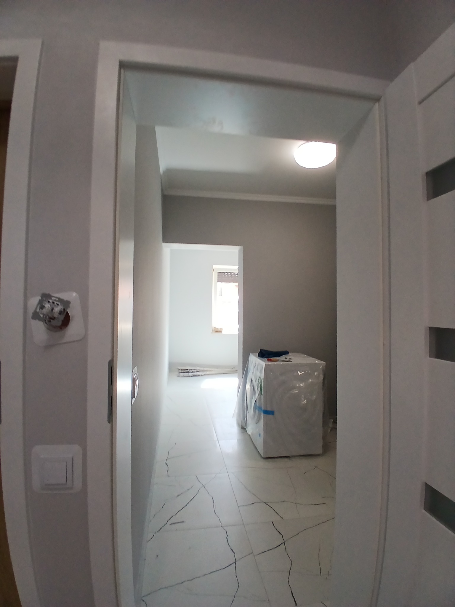 Wykończone wnętrze łazienki z marmurową podłogą i nową pralką w folii, widoczne przez białe drzwi. Świeżo pomalowane ściany w odcieniach szarości.