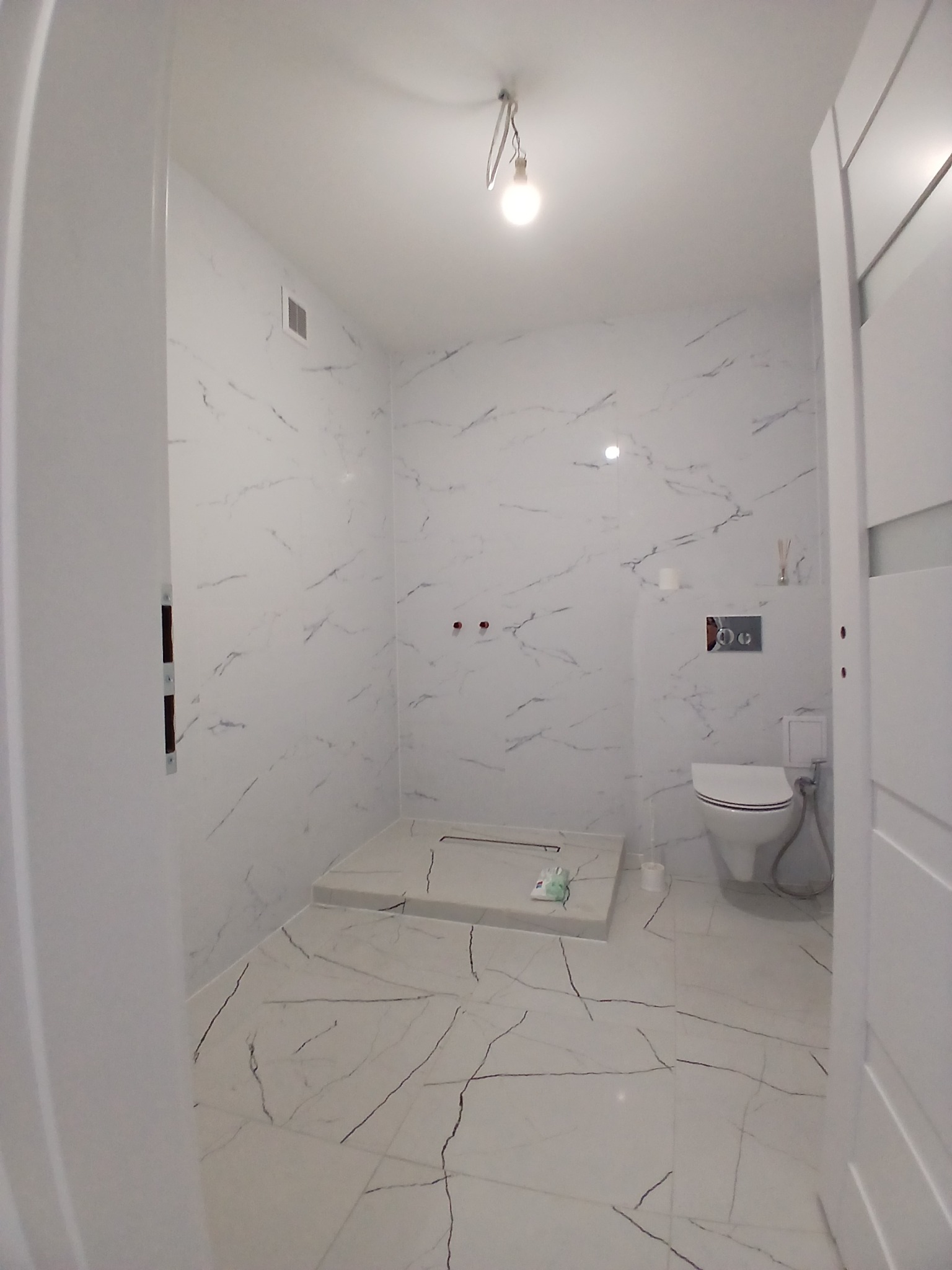 Wykończona łazienka w bieli z marmurkowym wzorem na płytkach. Widoczny brodzik bez kabiny, toaleta i minimalistyczne oświetlenie. Nowoczesny design.