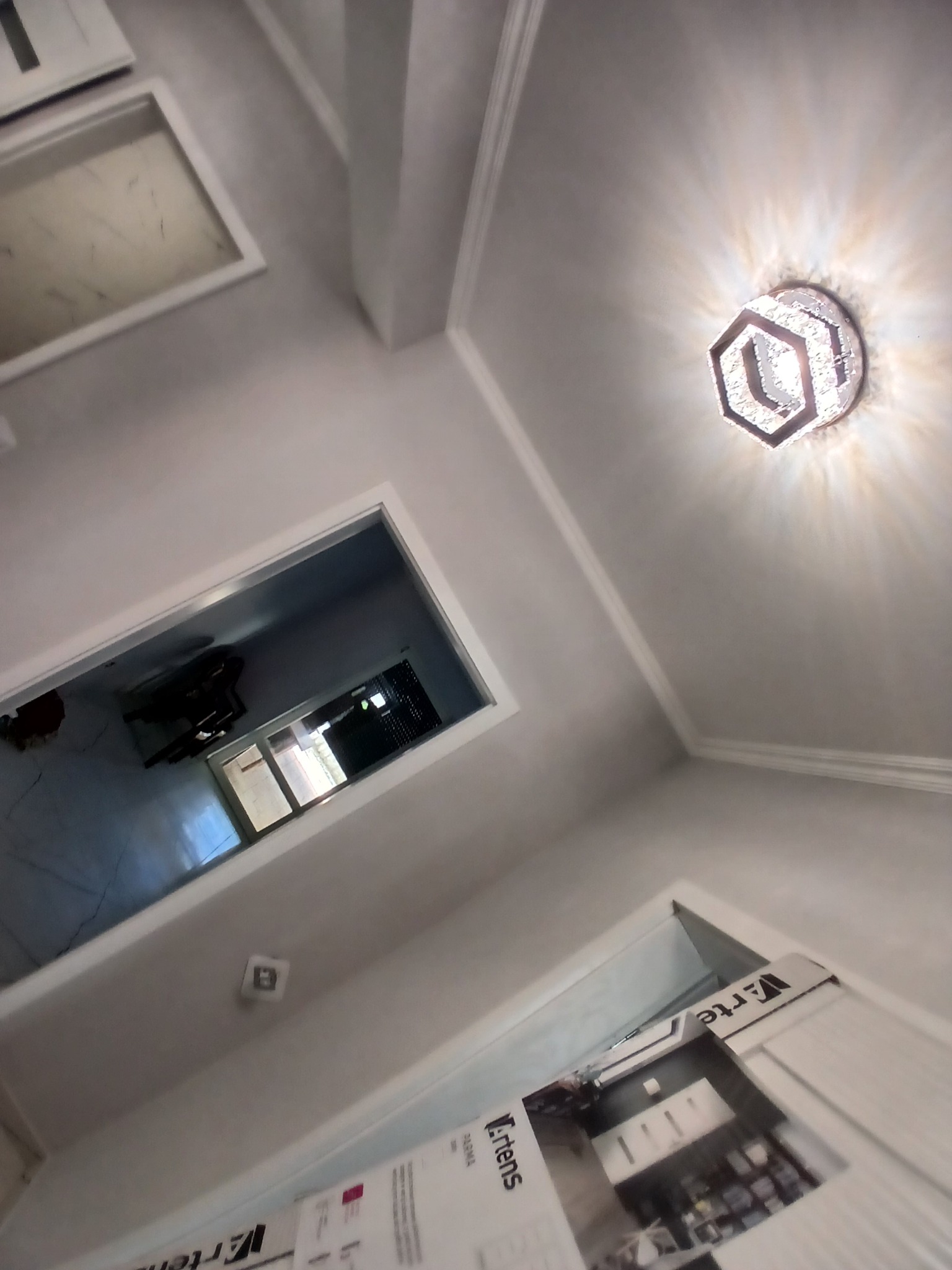 Nowoczesne wnętrze z geometryczną lampą sufitową, świeżo pomalowane ściany w odcieniach szarości i białe listwy dekoracyjne. Widok na okno i drzwi.