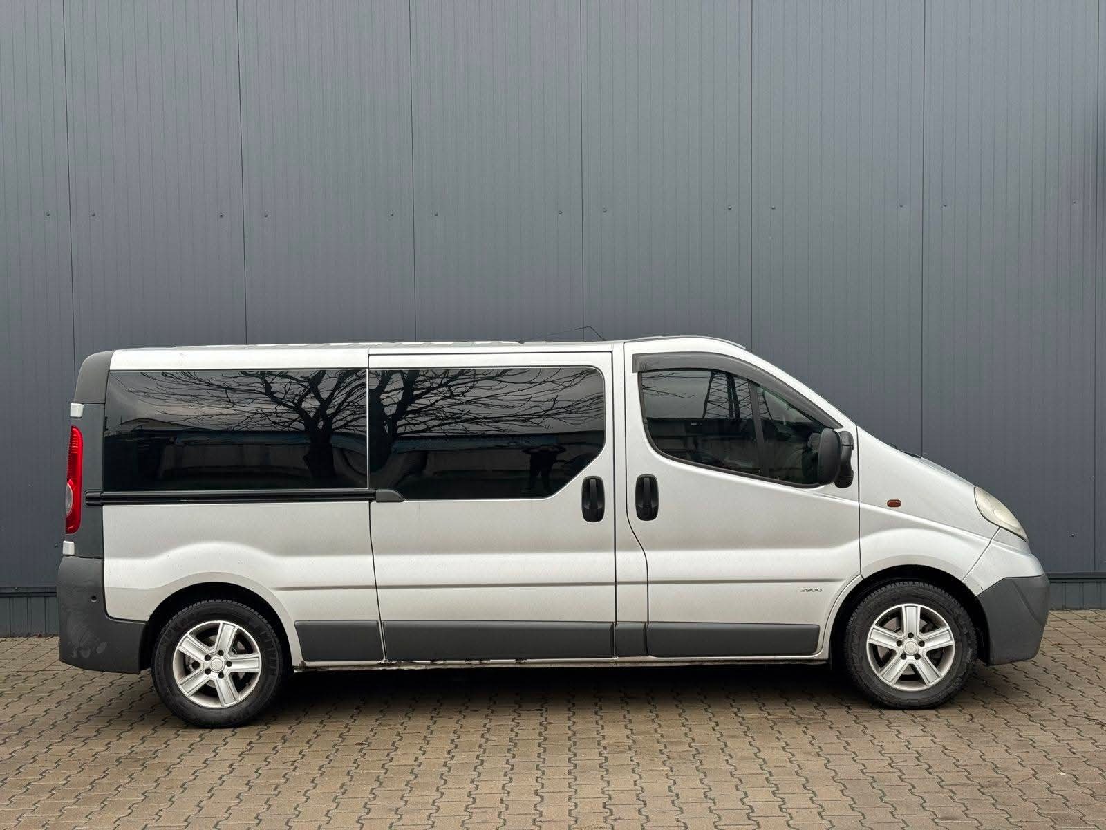 Srebrny bus z przyciemnianymi szybami, na których odbija się korona drzewa, zaparkowany na brukowanej nawierzchni na tle szarej ściany.