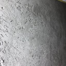 Szara, teksturowana ściana po szpachlowaniu z widoczną taśmą malarską przy suficie. Nieregularna faktura, światłocień podkreśla nierówności. Detal wykończenia wnętrza.