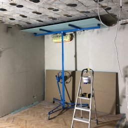 Montaż płyty G-K na stelażu w pomieszczeniu z surowym sufitem i parkietem. Widoczna drabina, narzędzia i płyty. Etap prac wykończeniowych.