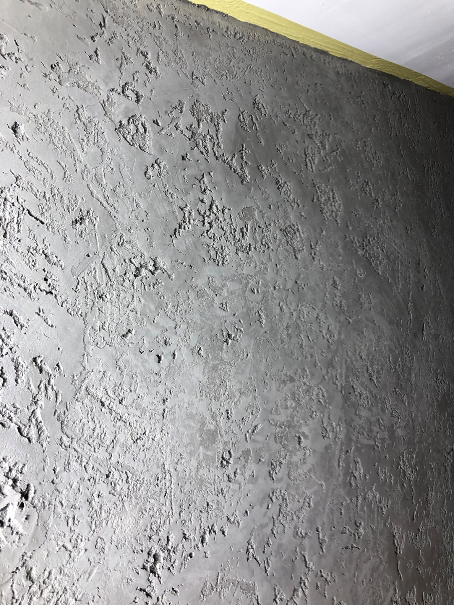 Szara, teksturowana ściana po szpachlowaniu z widoczną taśmą malarską przy suficie. Nieregularna faktura, światłocień podkreśla nierówności. Detal wykończenia wnętrza.