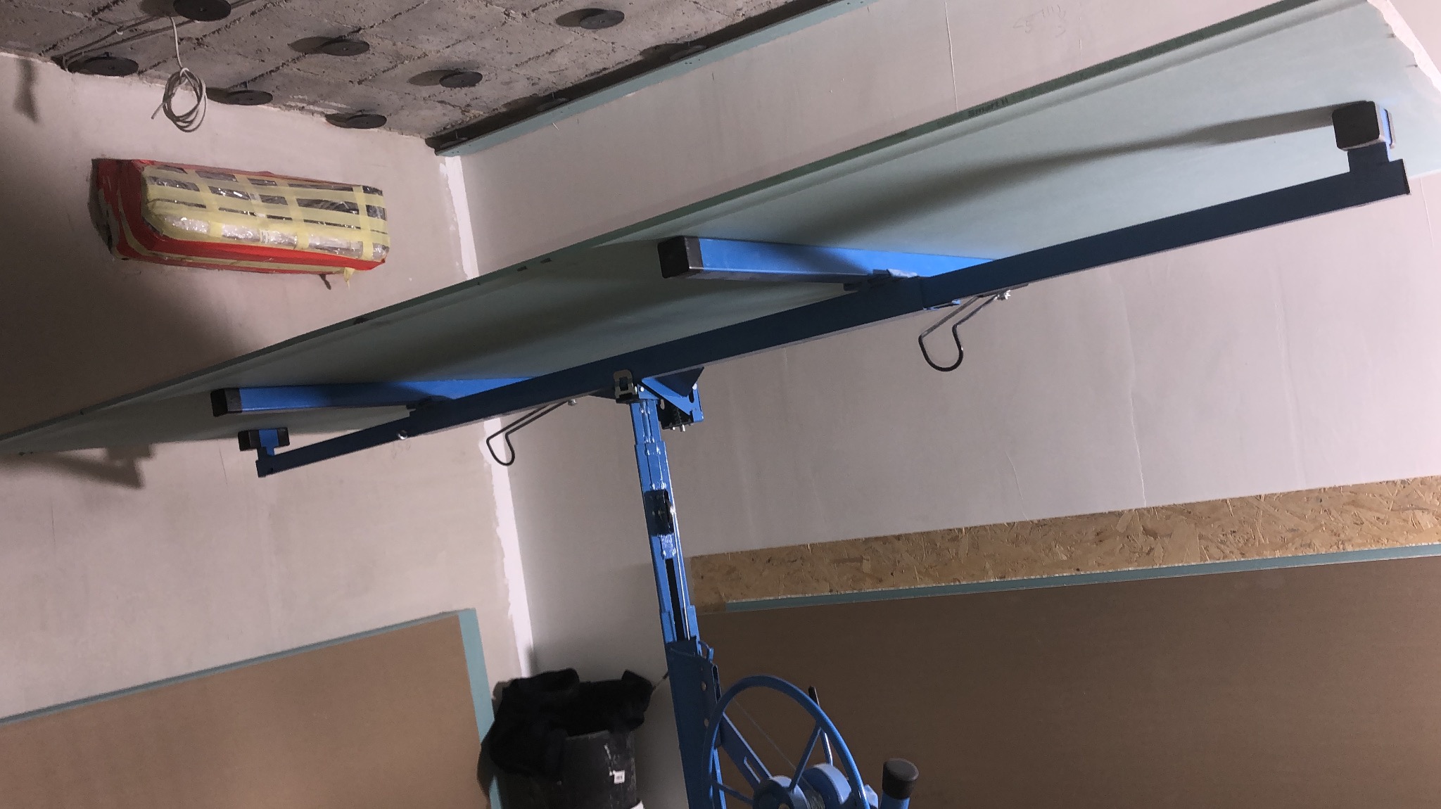 Montaż płyty G-K na suficie za pomocą podnośnika. Widoczny zabezpieczony klimatyzator i surowy stan ścian. Prace wykończeniowe w trakcie.