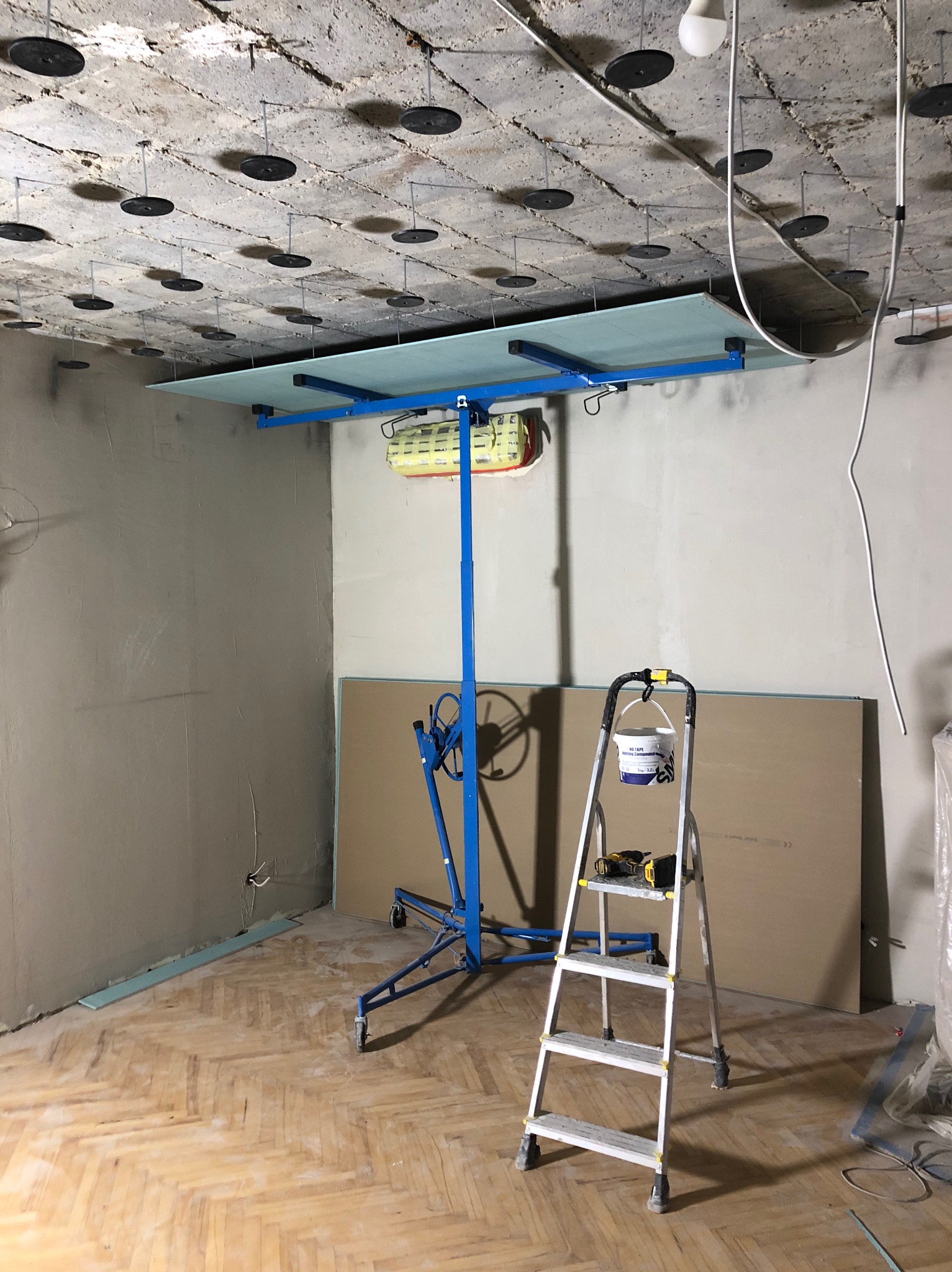 Montaż płyty G-K na stelażu w pomieszczeniu z surowym sufitem i parkietem. Widoczna drabina, narzędzia i płyty. Etap prac wykończeniowych.